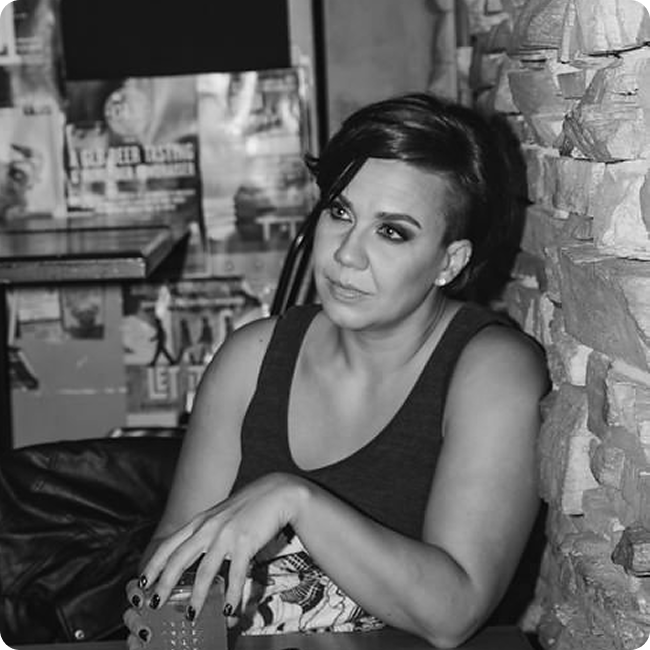 Black and white photo of a woman with short dark hair, wearing a sleeveless top, sitting at a table in a restaurant or cafe, with a brick wall behind her.