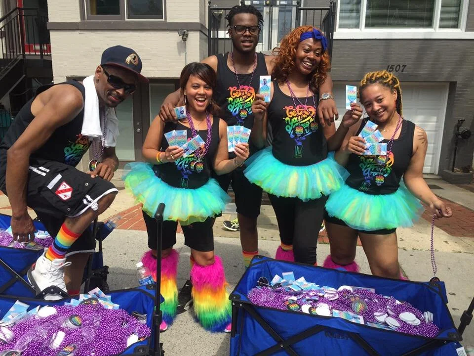 Group of five diverse people dressed in colorful costumes, holding Mardi Gras beads and tickets, standing outside in front of houses, with celebration decorations and bead-filled bins in front of them.
