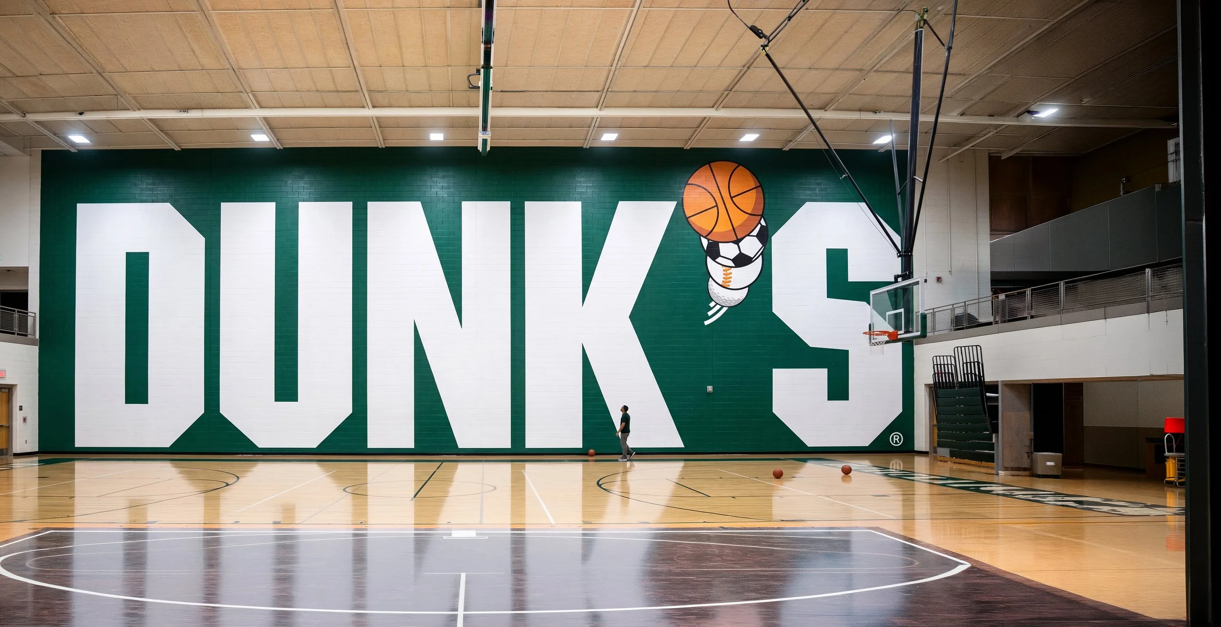 An indoor basketball court with a large wall featuring the word 'DUNK' in white letters, a basketball graphic, and a person shooting basketballs.