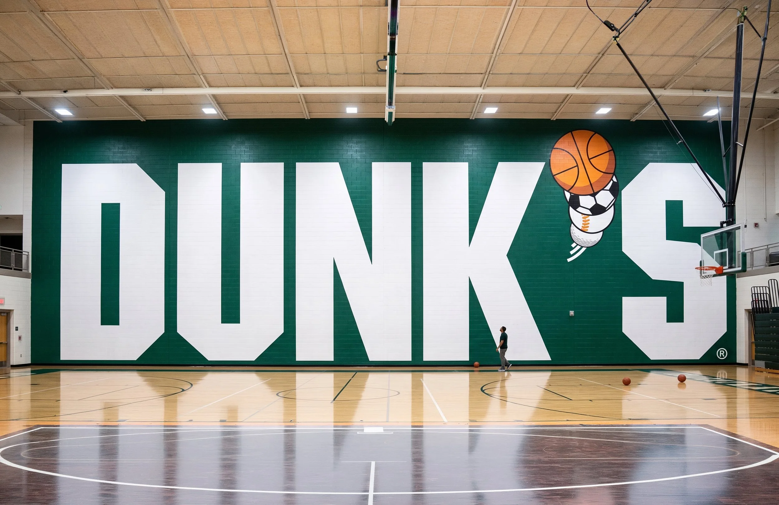 An indoor basketball court with a green wall featuring large white letters spelling 'THANKS' and a basketball illustration. There are two people near the wall and basketballs on the floor.