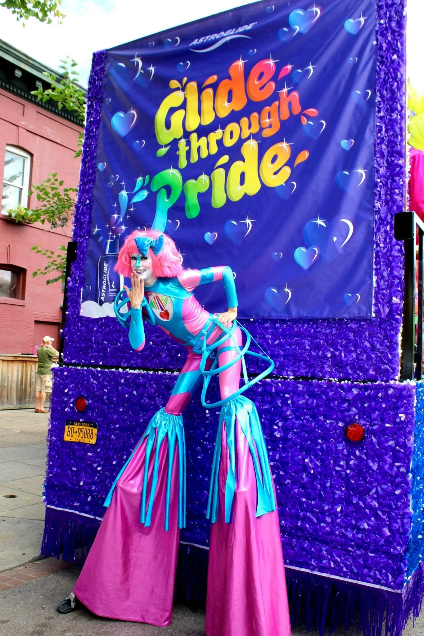 A person dressed as a colorful clown posing in front of a vibrant parade float decorated with purple and blue sequins, featuring a large banner that reads 'Guide Through Pride' with rainbow-colored text and hearts.