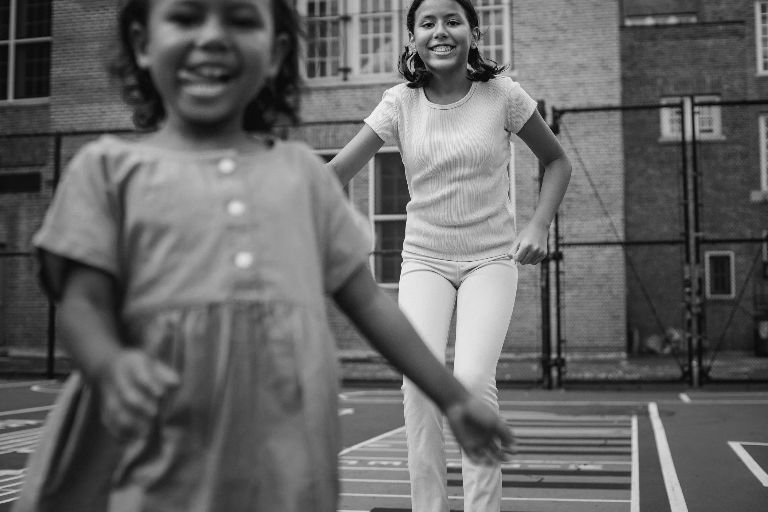 Two young girls playing in a basketball court, smiling and running. One girl in the foreground, blurred, and one girl in the background, clearly visible and smiling. The scene is outdoors with a brick building in the background.
