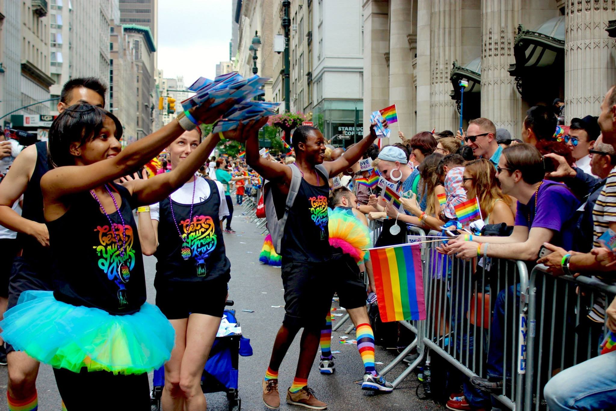 People participating in a Pride parade, wearing colorful outfits, holding rainbow flags, and cheering along a city street.