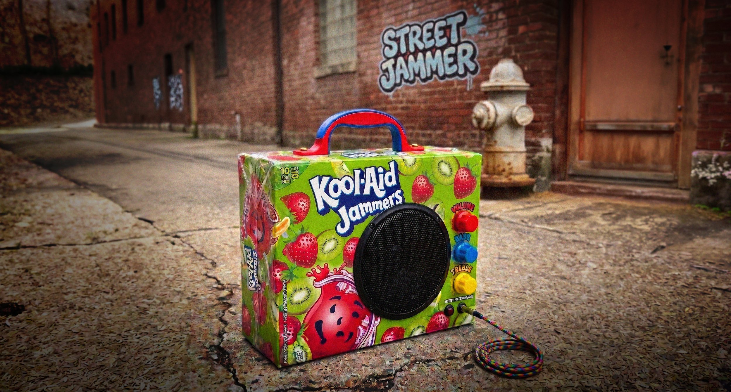 A colorful toy boombox with a handle, decorated with images of strawberries, kiwi, and watermelon on a city sidewalk. The boombox features large black speaker, red, blue, and yellow buttons, and a rainbow-colored cord. In the background, there is a brick building with graffiti that says 'Street Jammer', a fire hydrant, and a wooden door.