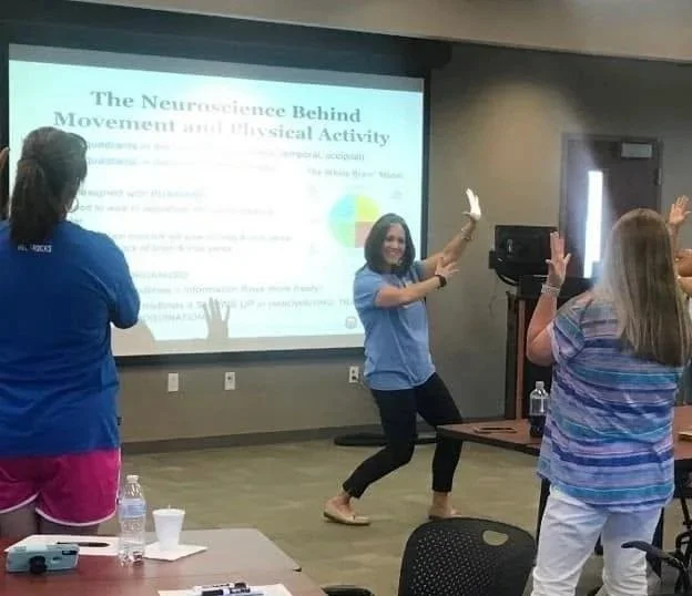 Dr. Stapp leading a dance activity in front of a presentation slide that reads 'The Neuroscience Behind Movement and Physical Activity.' She is smiling and moving her arms, with other women participating and raising their hands in the background.