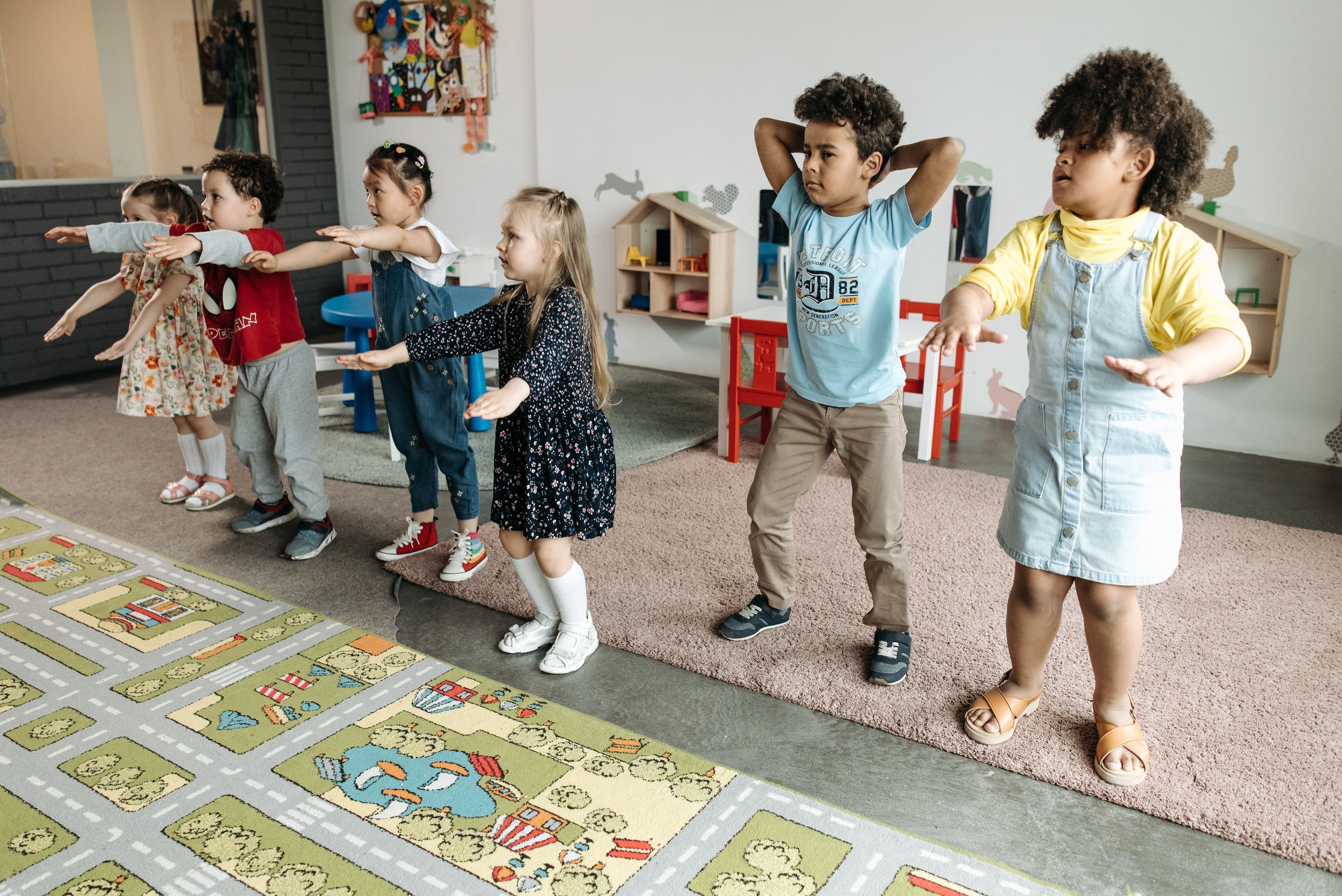 Group of young children standing on a carpet, participating in a classroom activity, with their arms extended forward in a line.