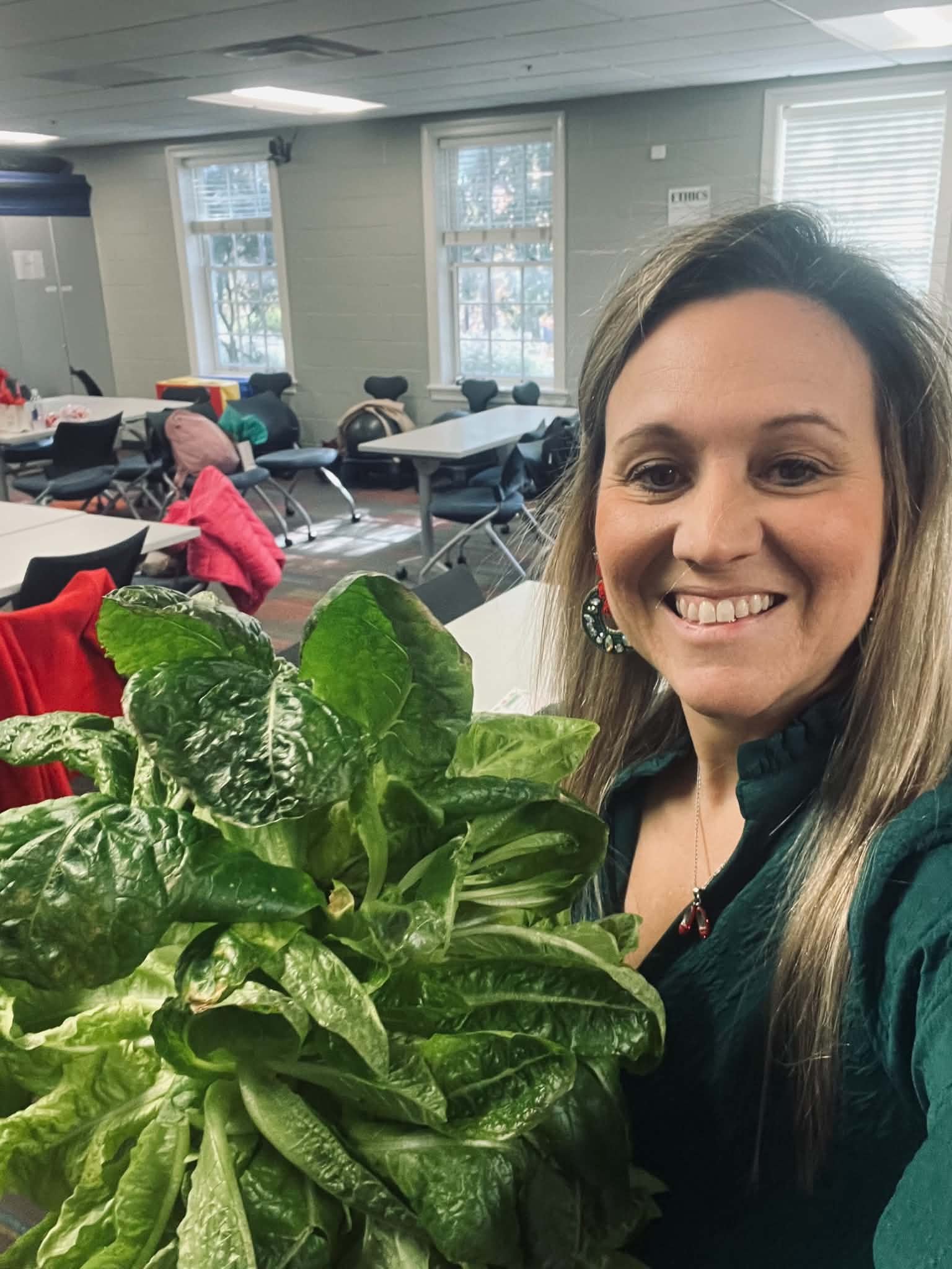 Dr. Stapp taking a selfie in a room with tables, chairs, and windows. She is smiling and has long, light brown hair with highlights. In the foreground, there is a large green leafy plant.