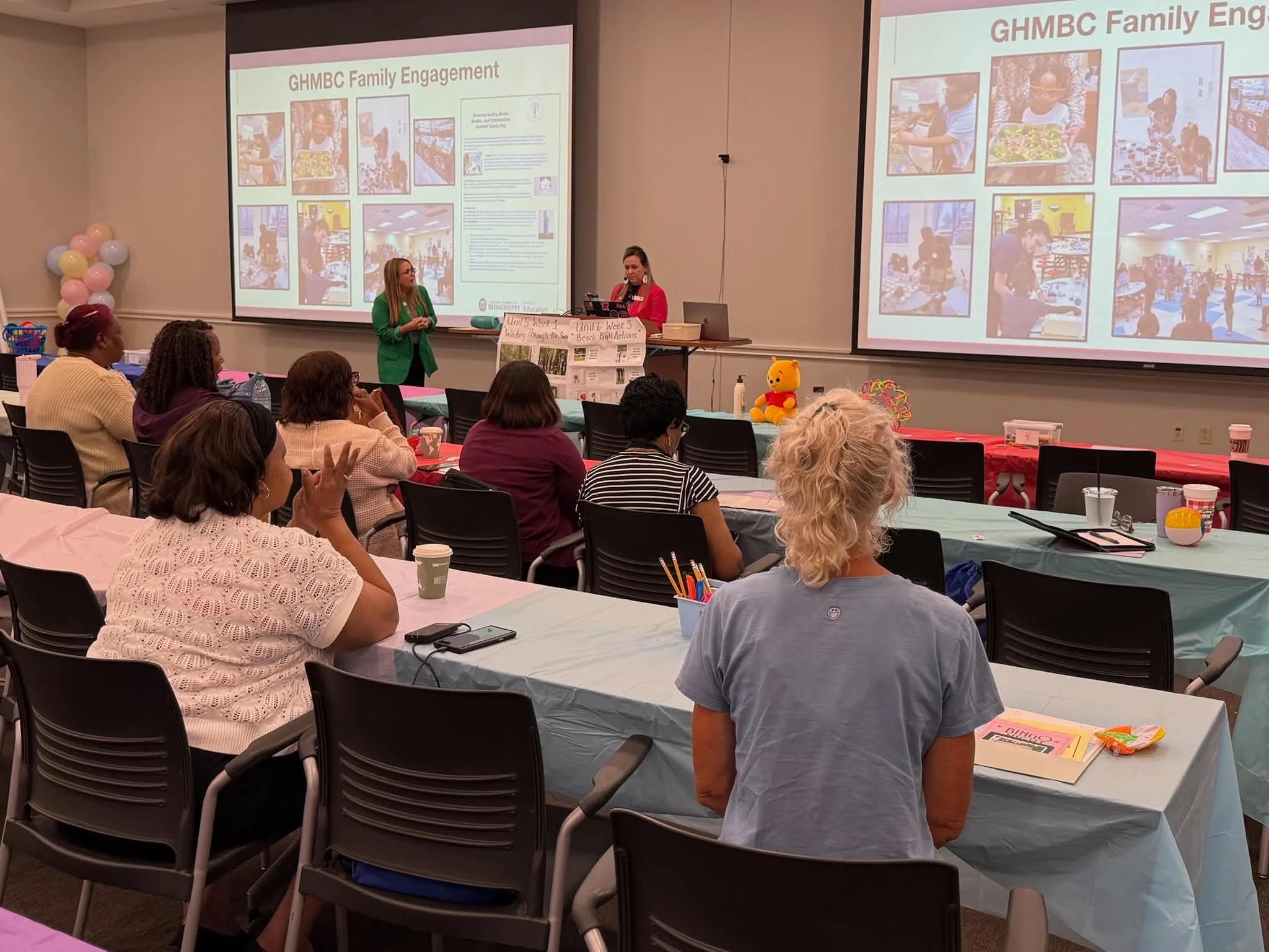 A conference room with several women of various ages seated at tables, watching a presentation with two large screens displaying slides about GHMBC Family Engagement. One woman in a red jacket is speaking, and another woman in a green jacket stands nearby. The tables are decorated with pink tablecloths and various items like coffee cups, a teddy bear, and flyers. There are balloons and a poster on the front table.