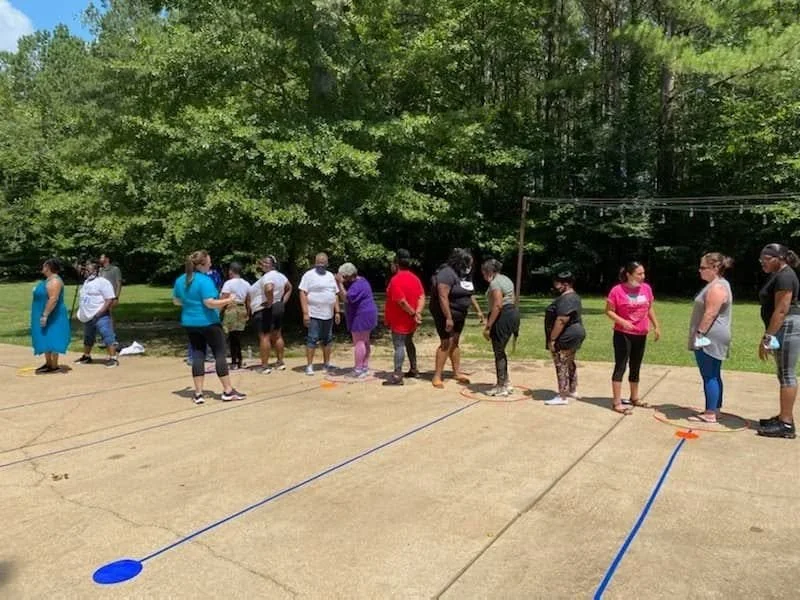 People standing in line outdoors on a sunny day, on a paved surface with colored lines and circles, near green trees.