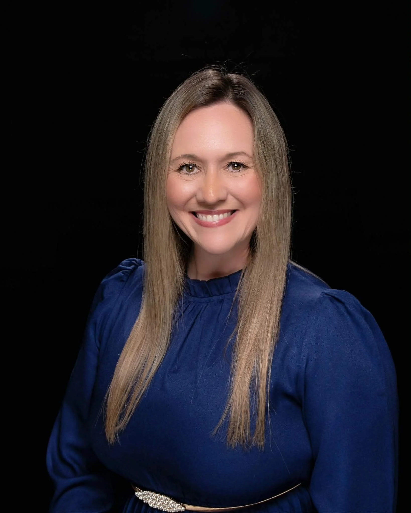 Portrait of Dr. Stapp with long straight blonde hair, wearing a dark blue dress with puffed sleeves and a pearl embellishment at the waist, against a black background.