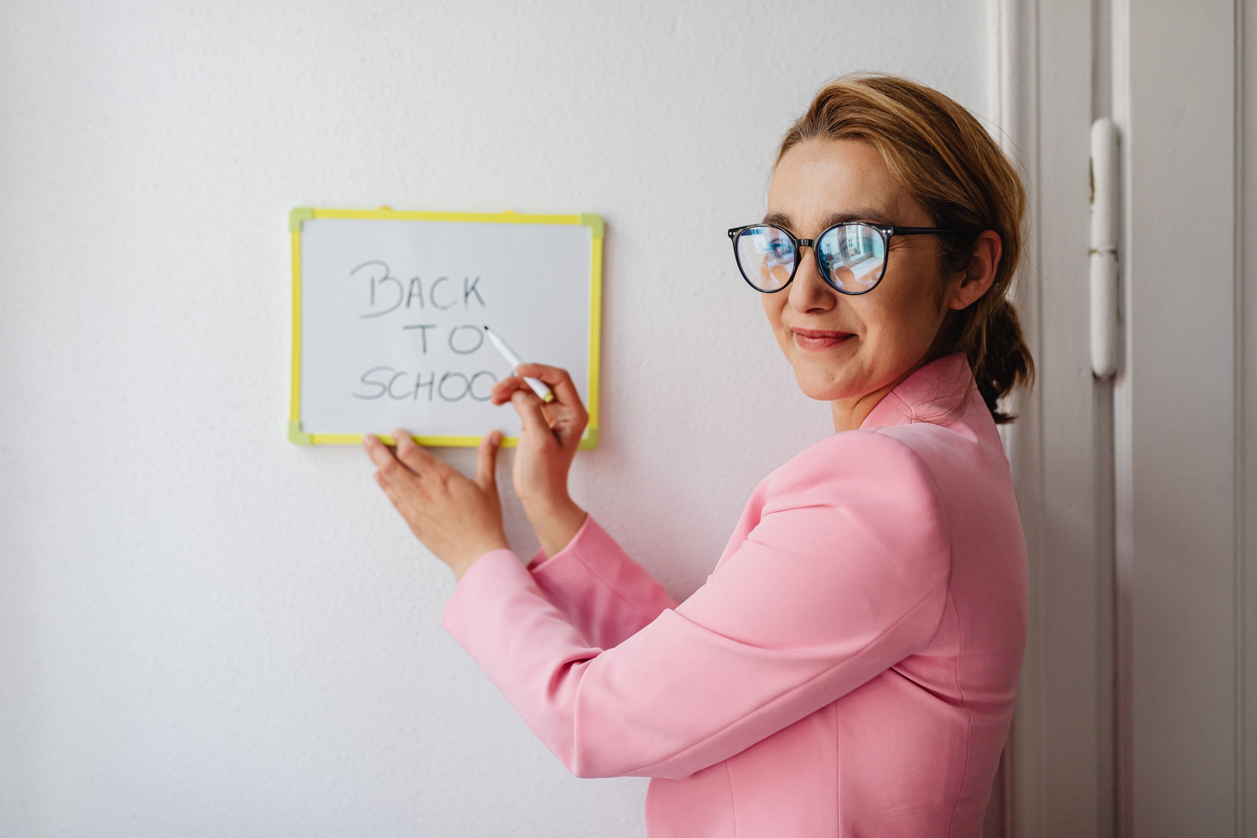 A woman with short brown hair, wearing glasses and a pink blazer, is writing on a whiteboard that says "BACK TO SCHOOL" in black marker. She is smiling and standing near a plain white wall.