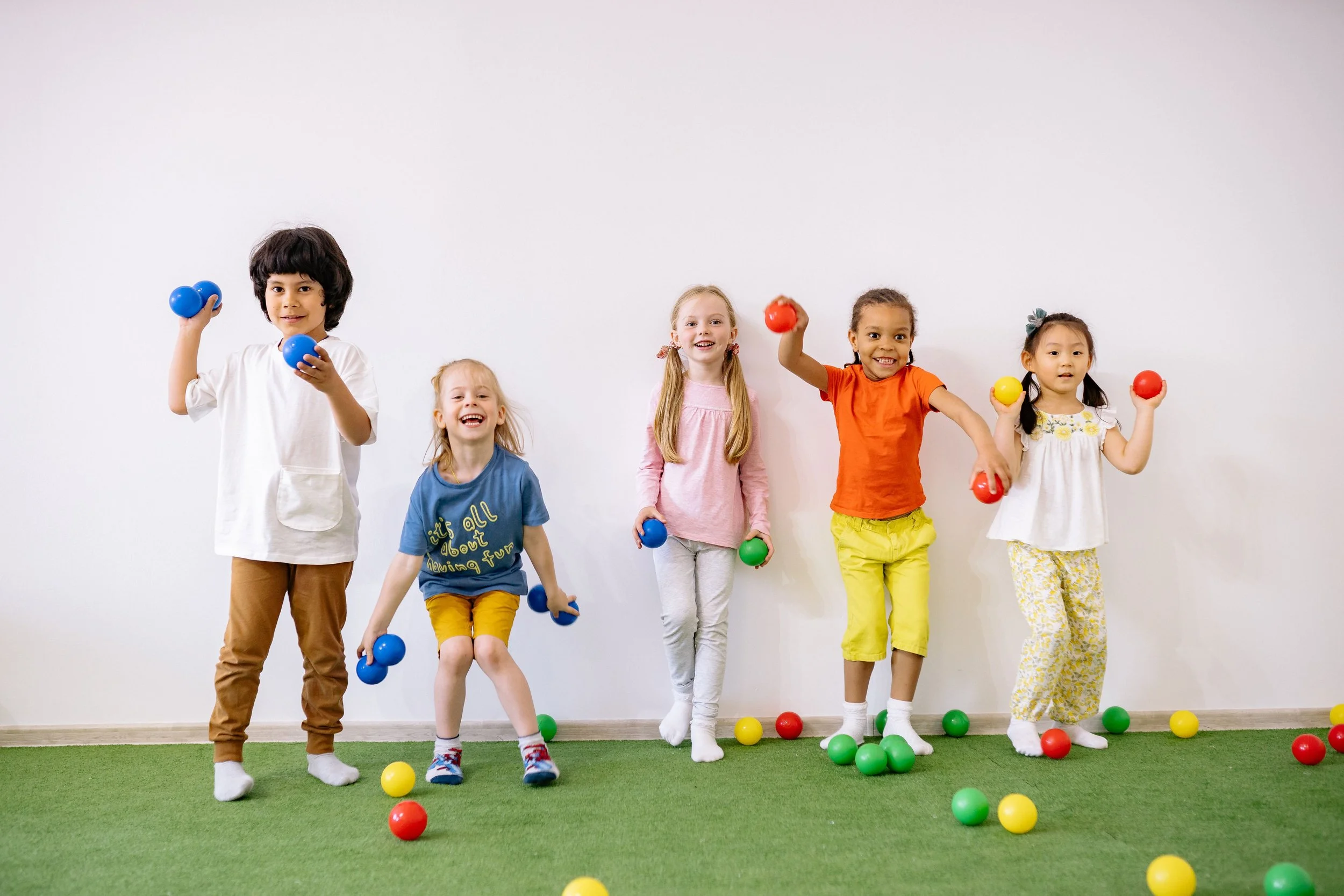 Six children standing on green artificial grass, playing with colorful balls in front of a plain white wall.