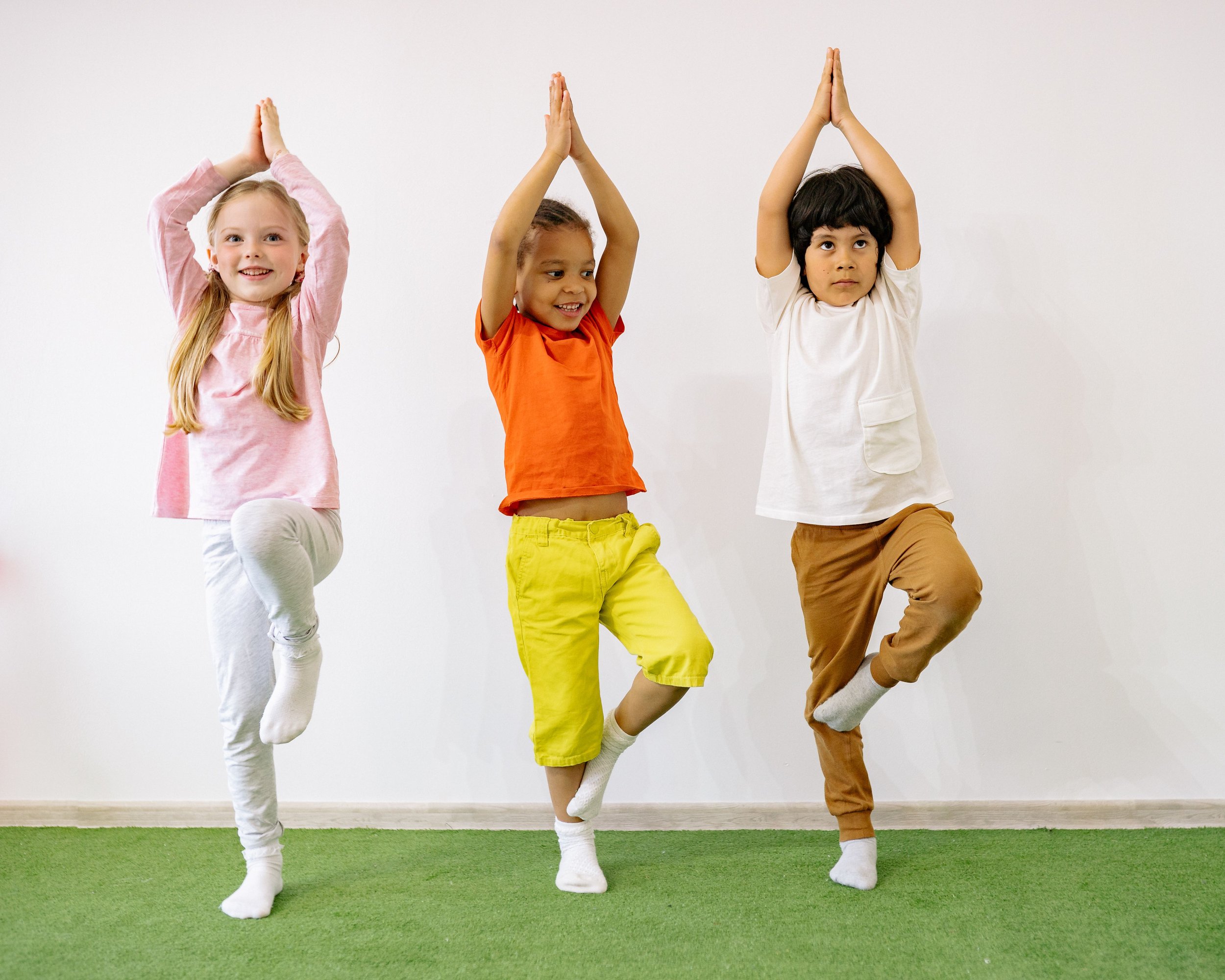 Three children standing on one leg, with arms raised above their heads, balancing on one foot inside a room with a white wall and green floor.