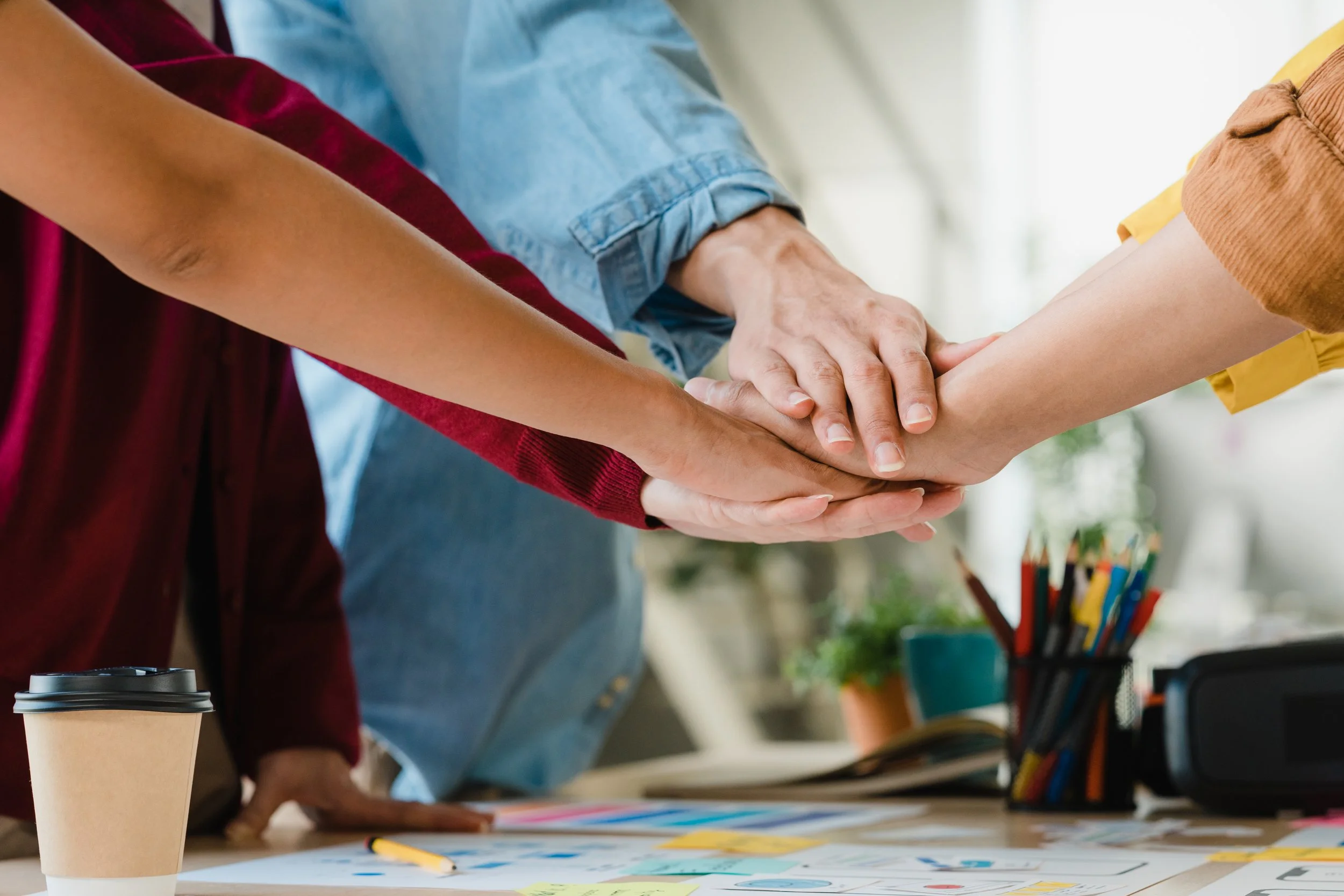 Three people with different skin tones placing their hands together over a table, likely in a team-building gesture. In the background, there are art supplies and a coffee cup on the table, indicating a creative or collaborative setting.