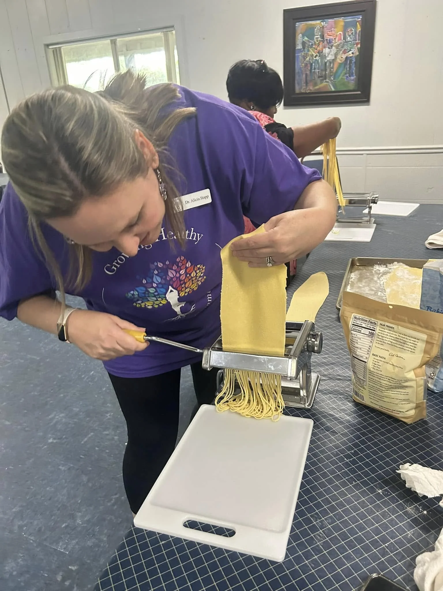 Two women preparing pasta using a manual pasta machine in a room with a black checkered tablecloth, a bag of pasta, and two white cutting boards.