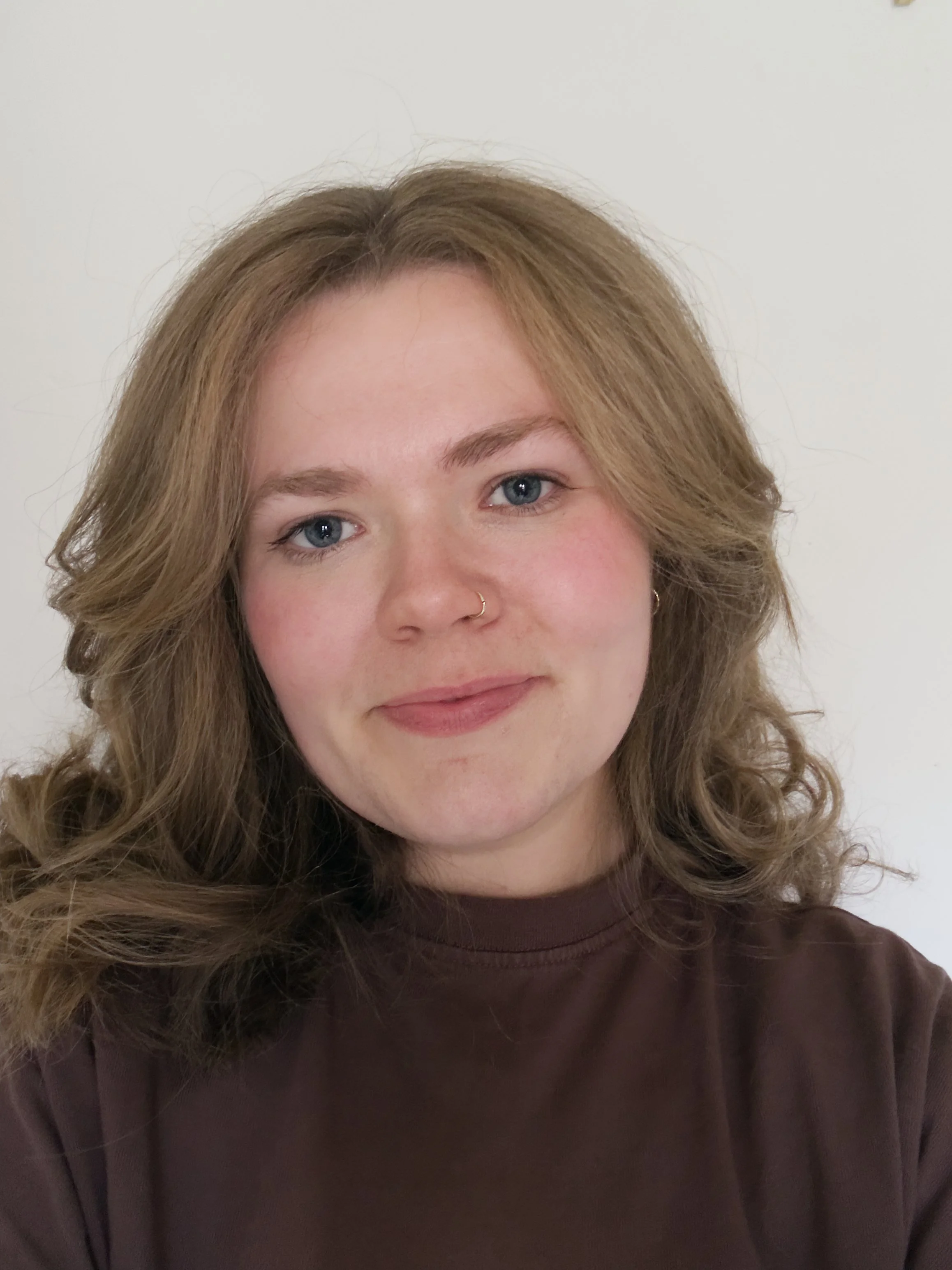 Close-up of a smiling woman with curly light brown hair and a nose ring, wearing a dark brown top against a plain white wall background.
