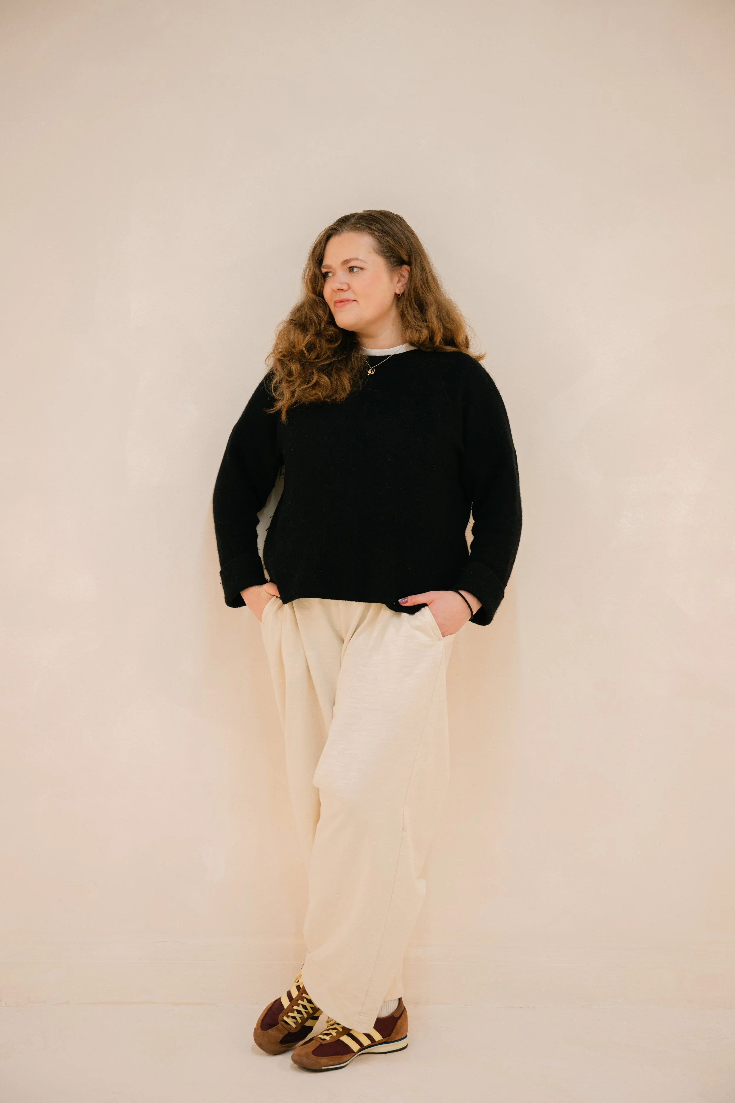 A woman with long curly hair wearing a black sweater, beige wide-leg pants, and multicolored sneakers standing against a plain off-white wall.