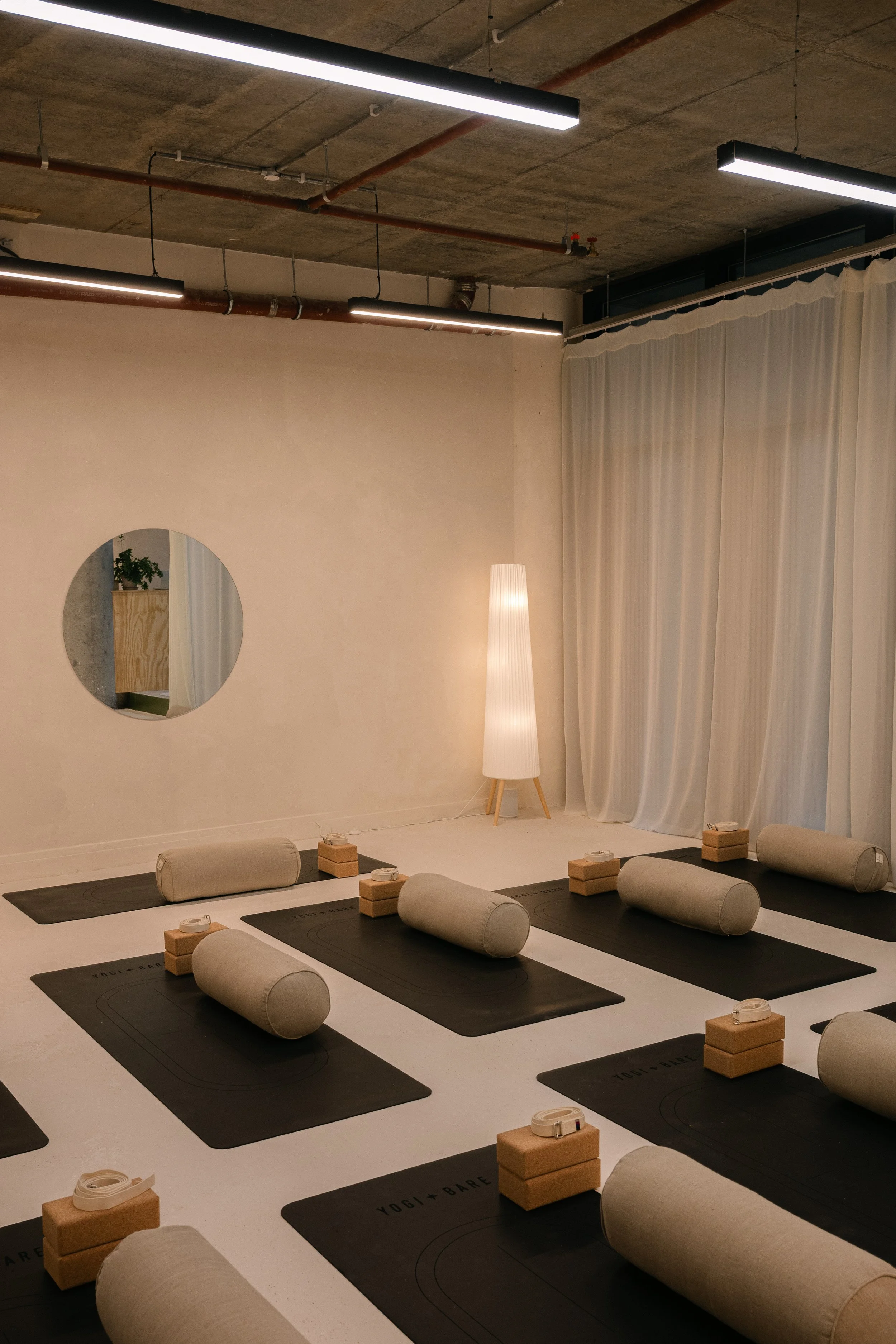 Yoga mats arranged in a classroom setting, each with a bolster, yoga blocks, and a strap, in a room with a concrete ceiling, long ceiling lights, and cream curtains.