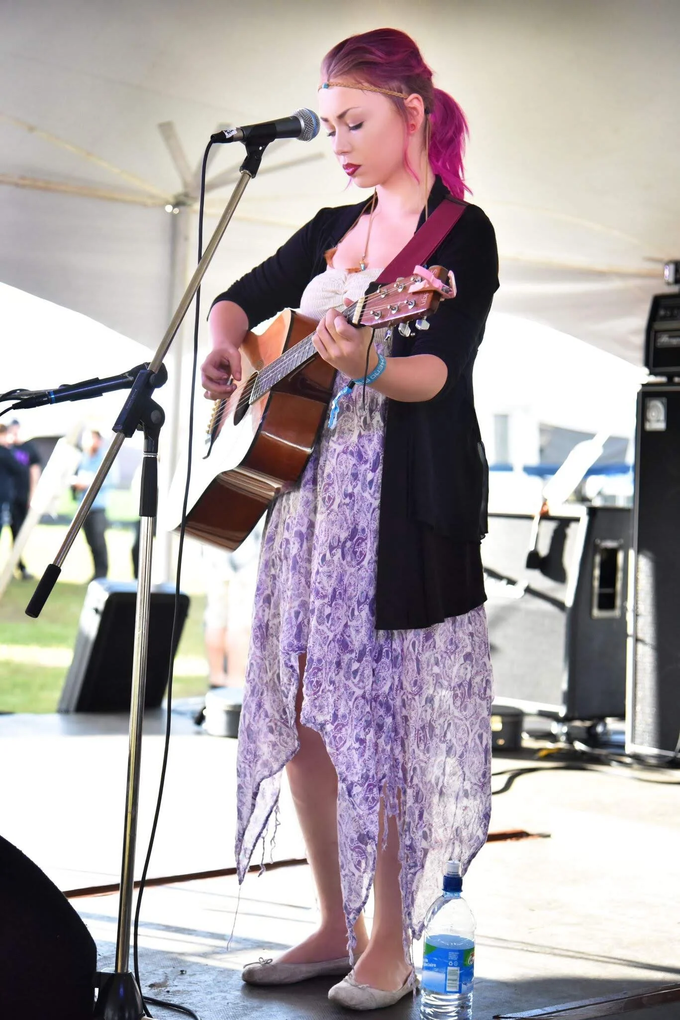 A young woman with pink hair singing into a microphone while playing an acoustic guitar on a stage. She is wearing a black cardigan, a light-colored dress, and flat shoes. There is a water bottle on the floor next to her, and audio equipment visible in the background.