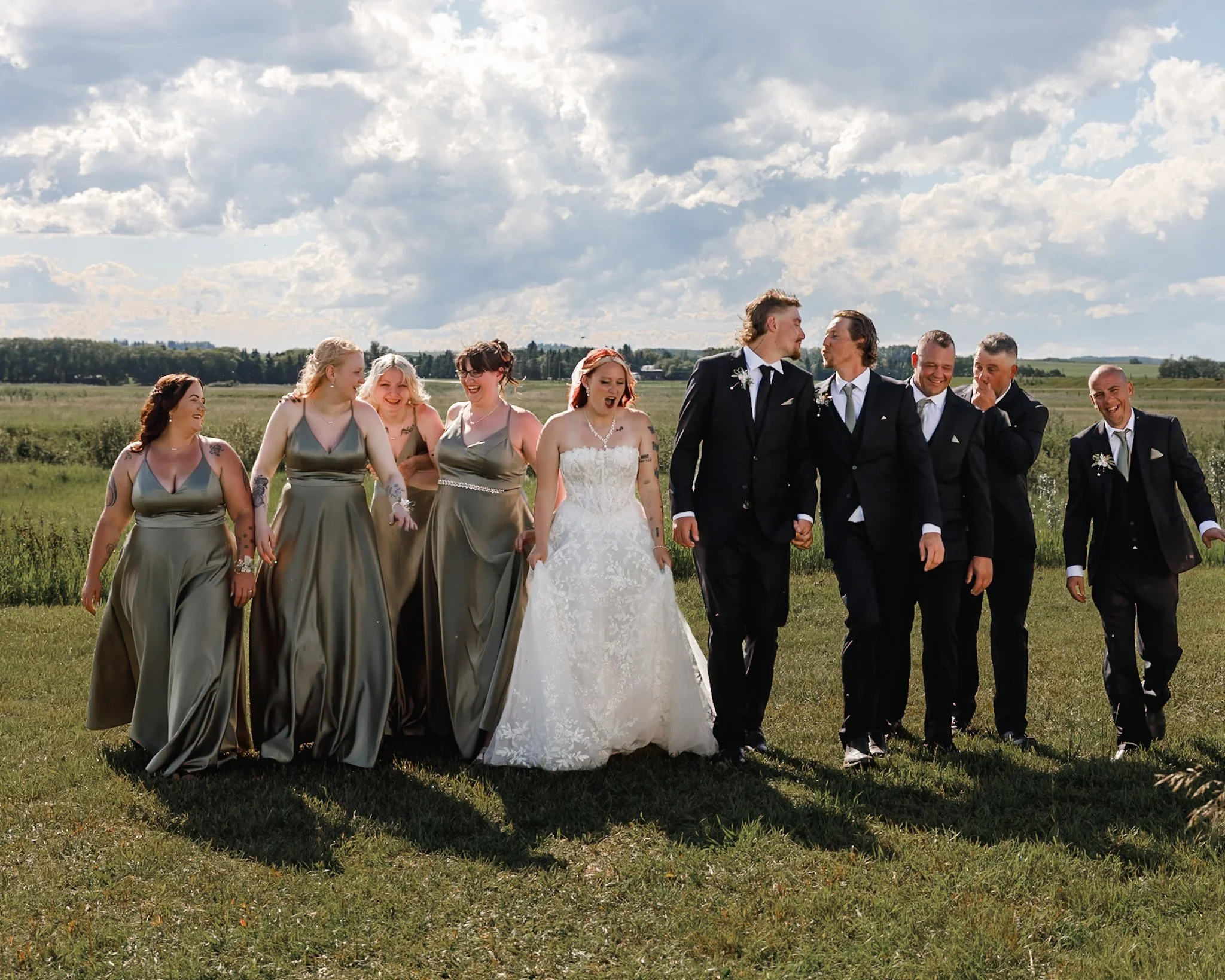 A group of men and women dressed in formal attire walking outdoors in a grassy field under a partly cloudy sky, appearing to be at a wedding celebration.