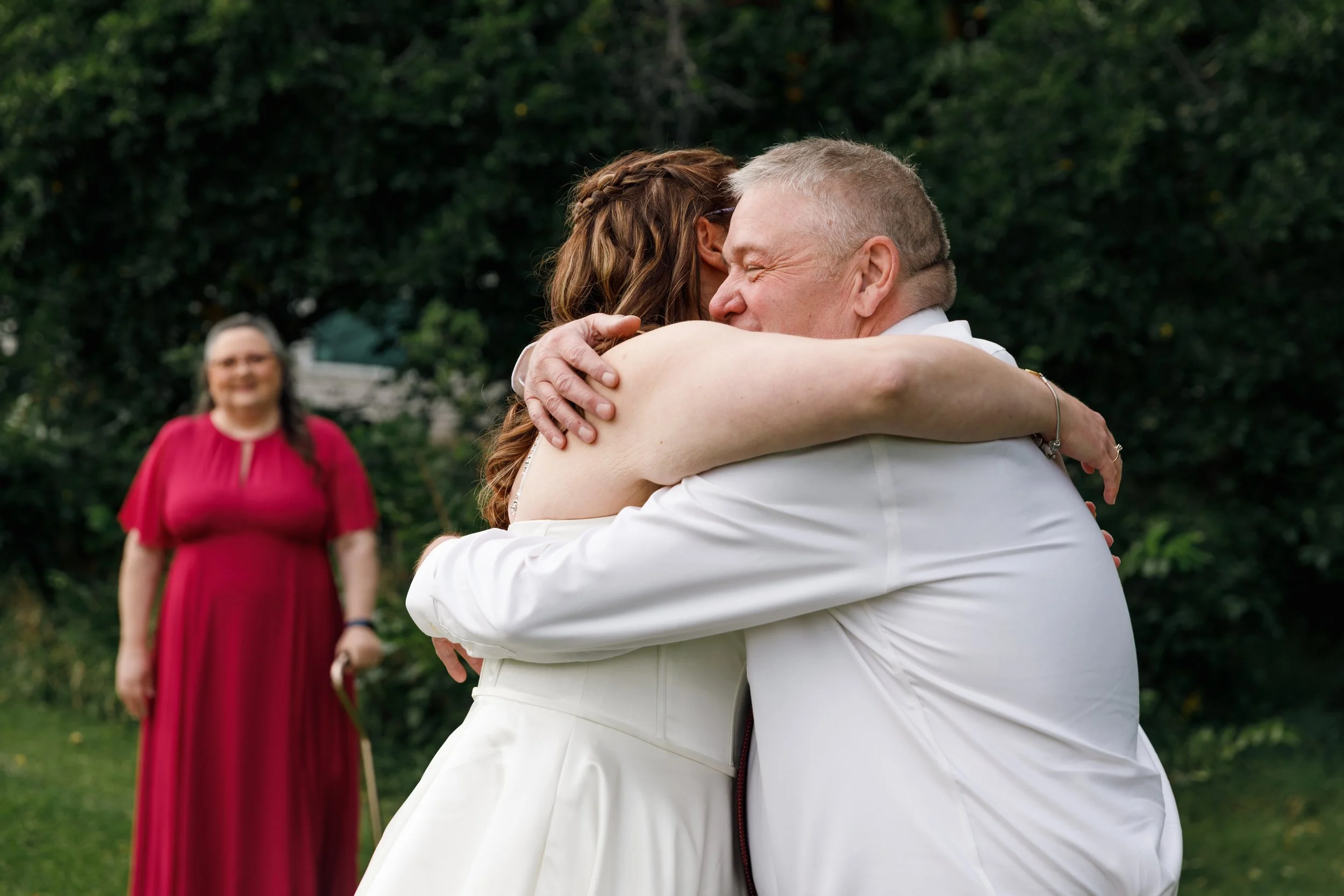 Two people hugging outdoors with greenery in the background, a woman in red standing and smiling in the distance.
