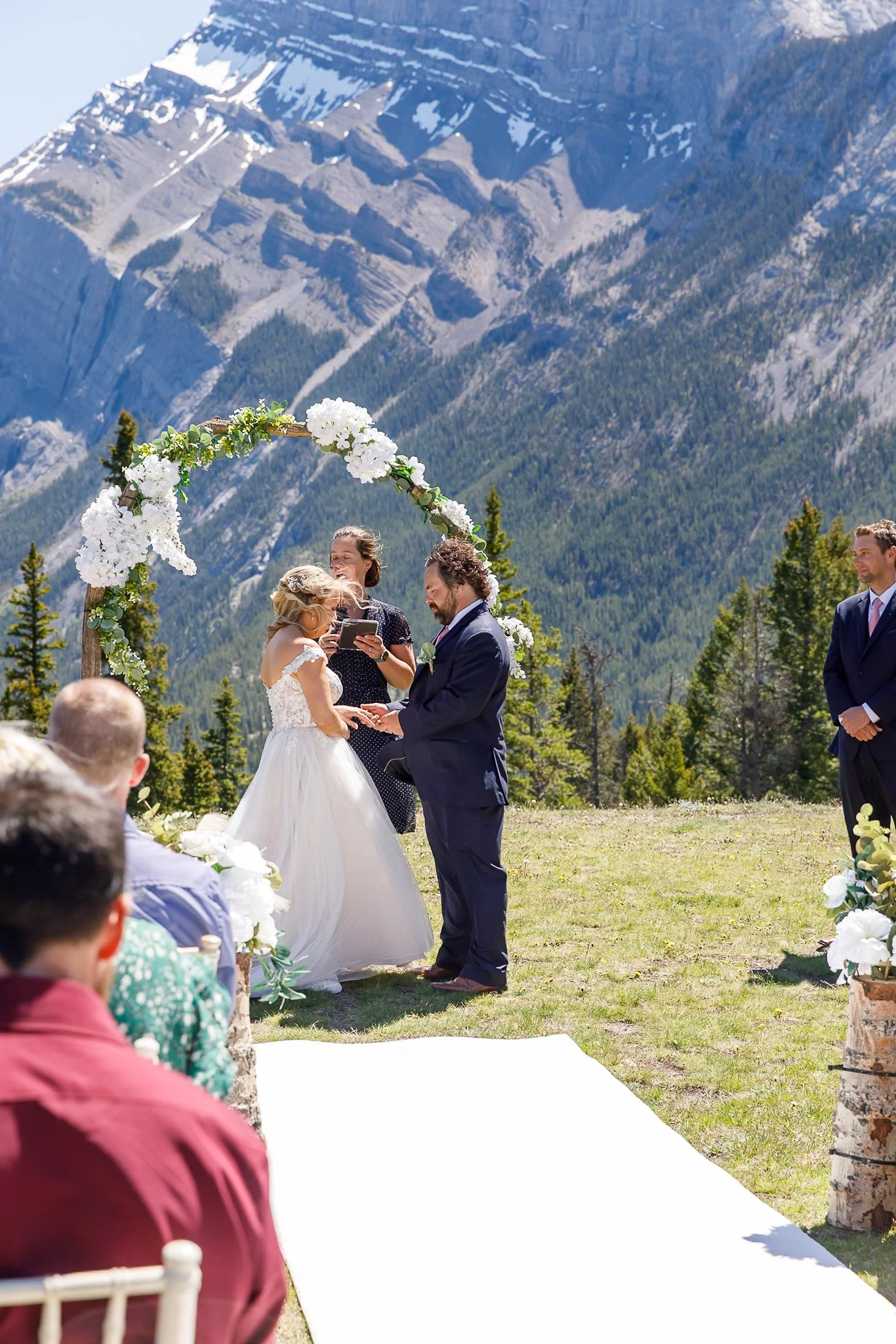 A wedding ceremony outdoors with a mountainous backdrop, a bride and groom exchanging vows under an arch decorated with white flowers, surrounded by seated guests.