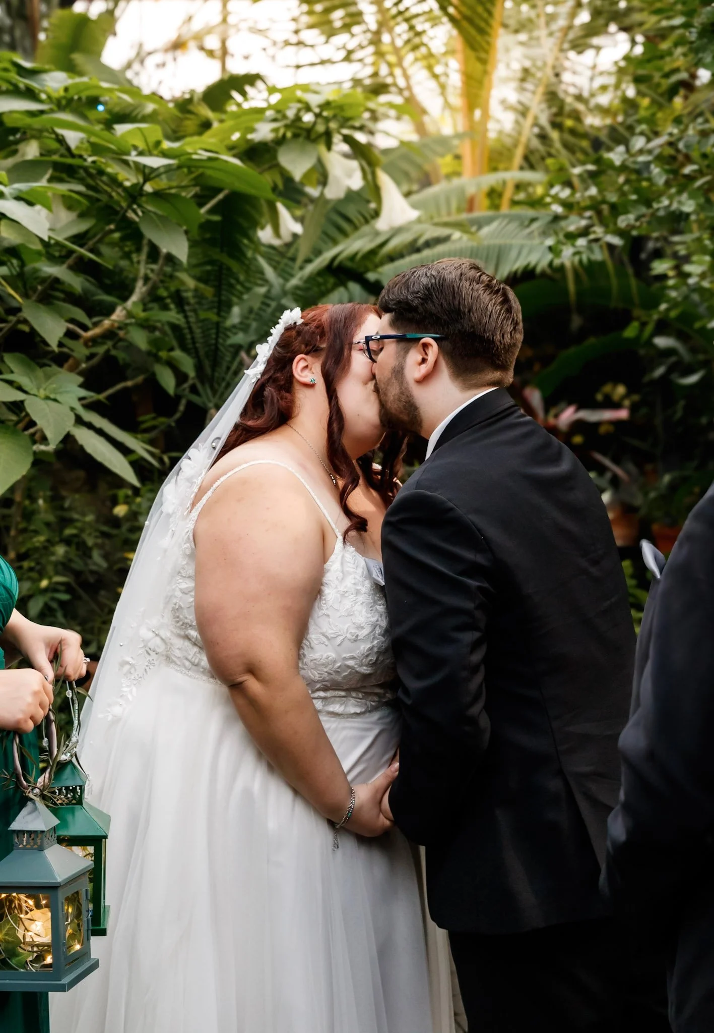 A bride and groom kissing during a wedding ceremony surrounded by lush green tropical plants.