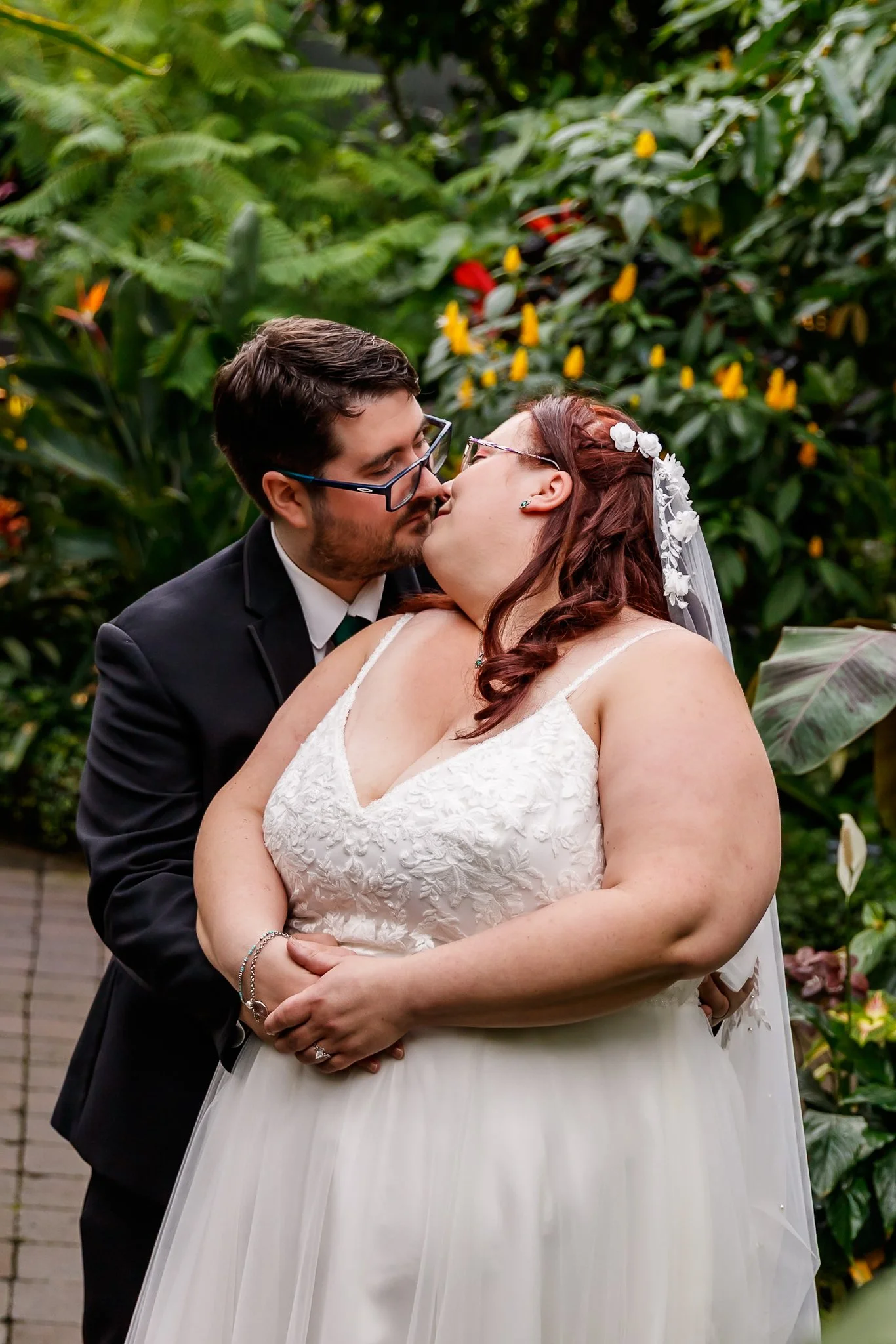 A bride and groom kissing outdoors surrounded by green plants, with the bride wearing a white lace wedding dress and the groom in a black suit and glasses.