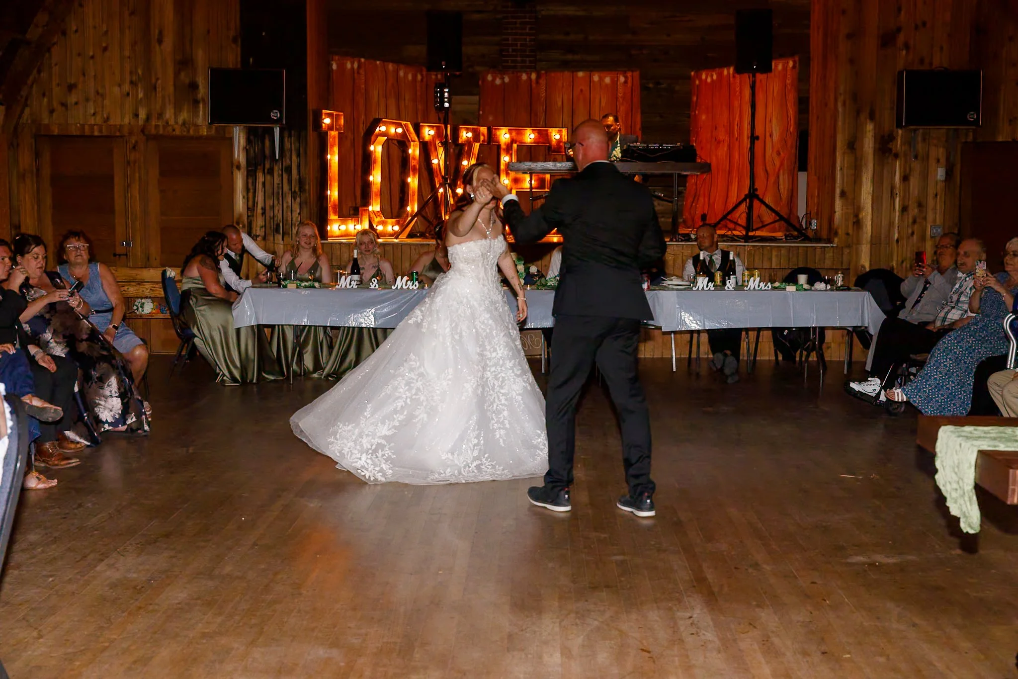 A bride and groom are dancing at a wedding reception in a rustic venue with wooden walls. Guests are seated at a long table watching and taking photos, with decorative 