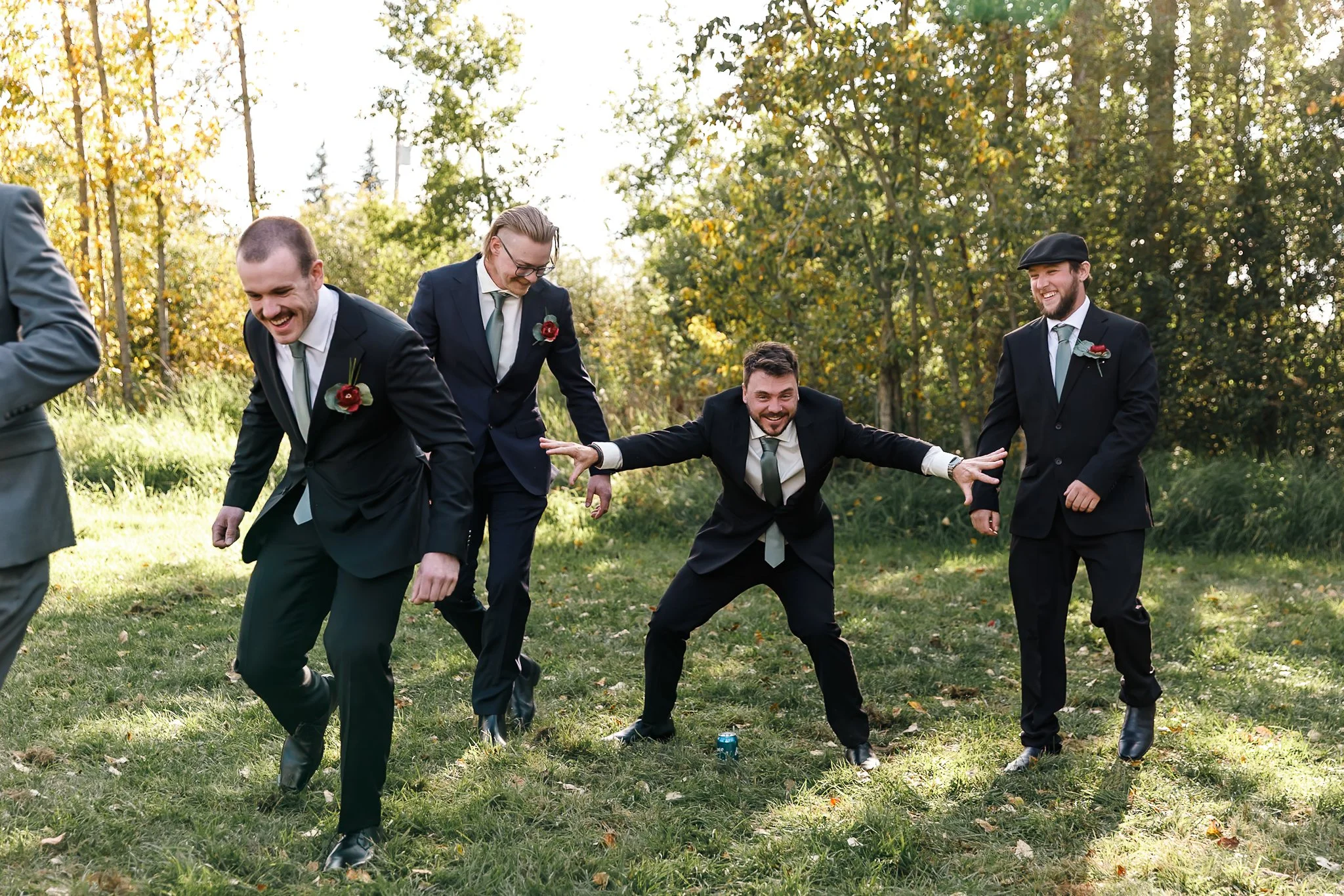 A group of five men in suits and ties, some wearing boutonnières, playing outdoors in a grassy area with trees. They are laughing and pose in a playful manner, with one man in the center squatting and spreading his arms, surrounded by others also smi