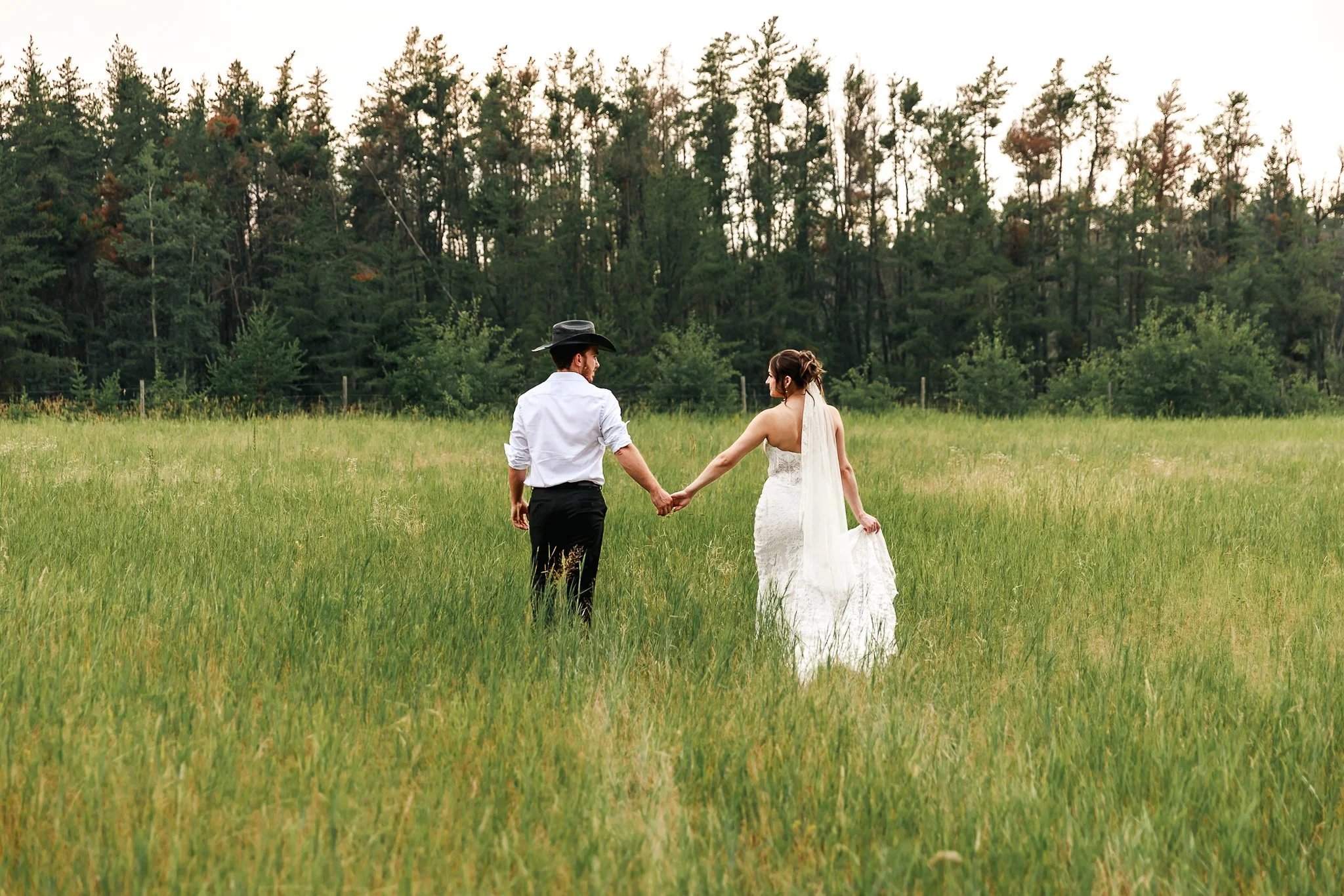 A couple holding hands walking through a grassy field near a wooded area, with the woman wearing a white wedding dress and the man wearing a light shirt and black hat.