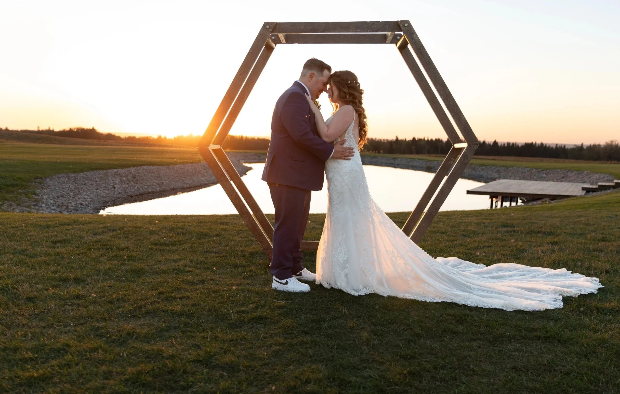 A newlywed couple is embracing and touching foreheads during sunset outdoors. The bride is in a white lace wedding gown with a long train, and the groom is in a dark suit. They are standing on grass near a body of water, with a wooden geometric arch framing them.