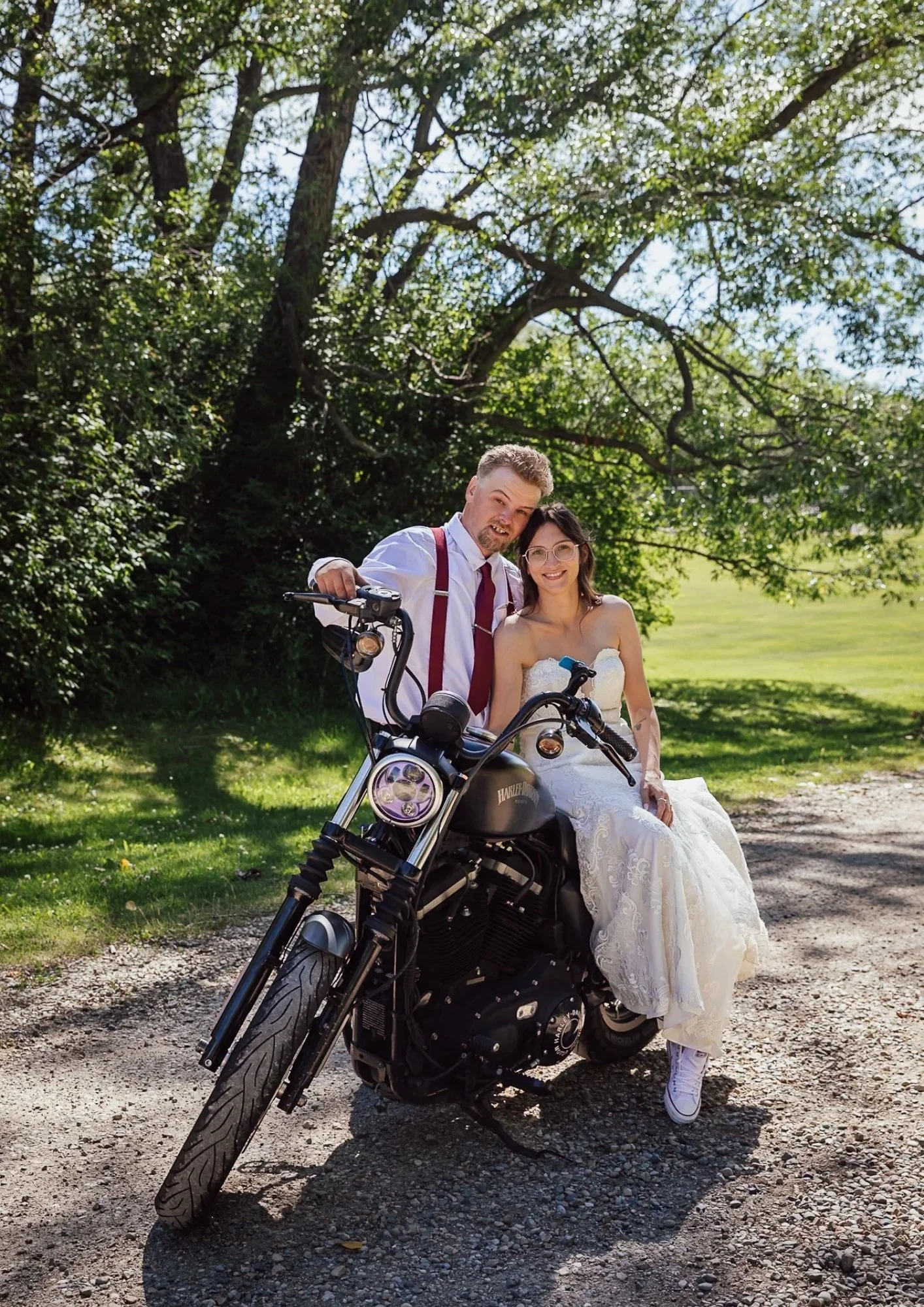A couple dressed in wedding attire sitting on a black motorcycle outdoors on a gravel path, with large trees and greenery in the background, sunny weather.