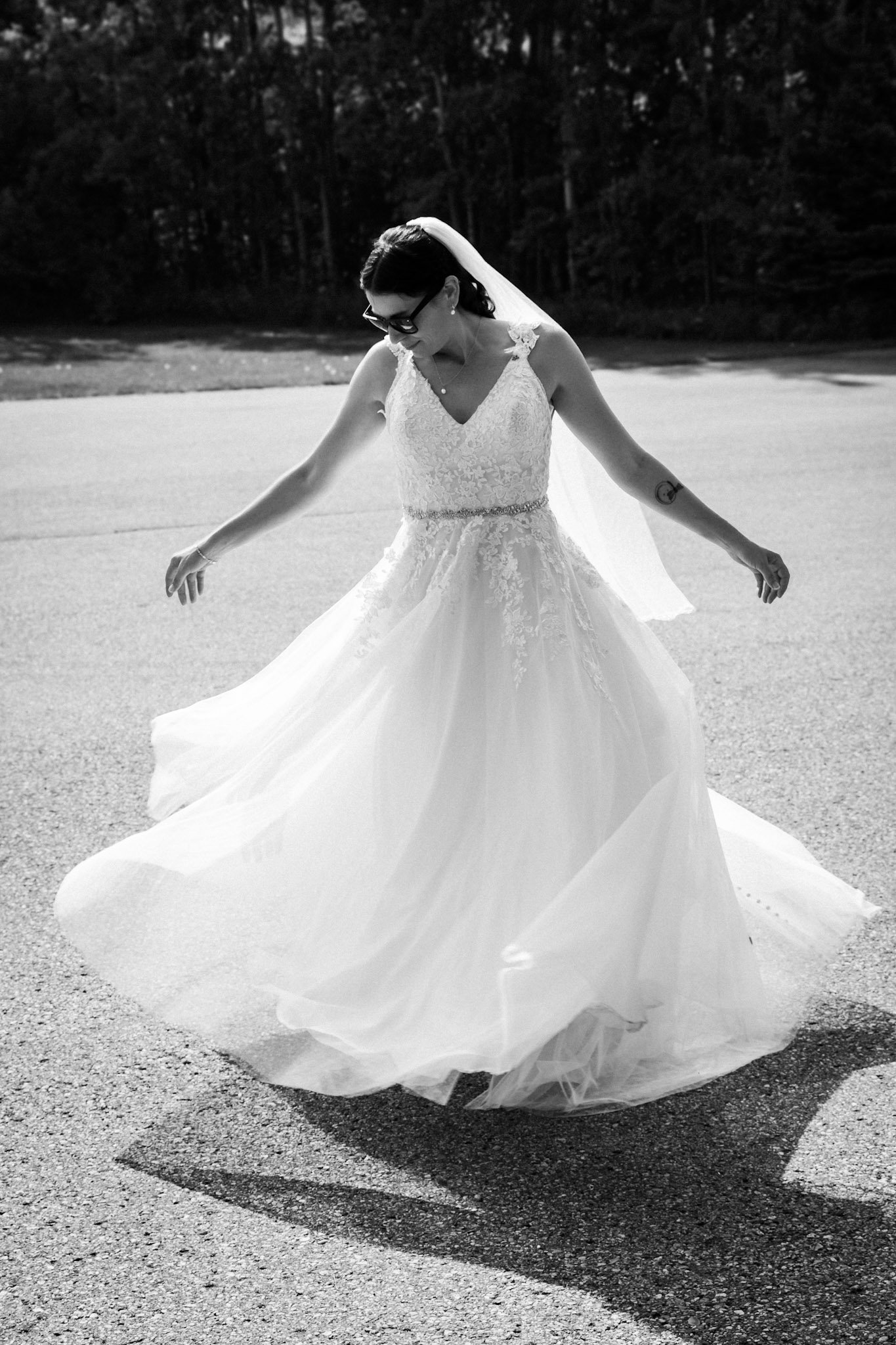 Black and white photo of a bride in a wedding dress spinning or dancing outdoors, with her dress flowing around her and her shadow visible on the ground.