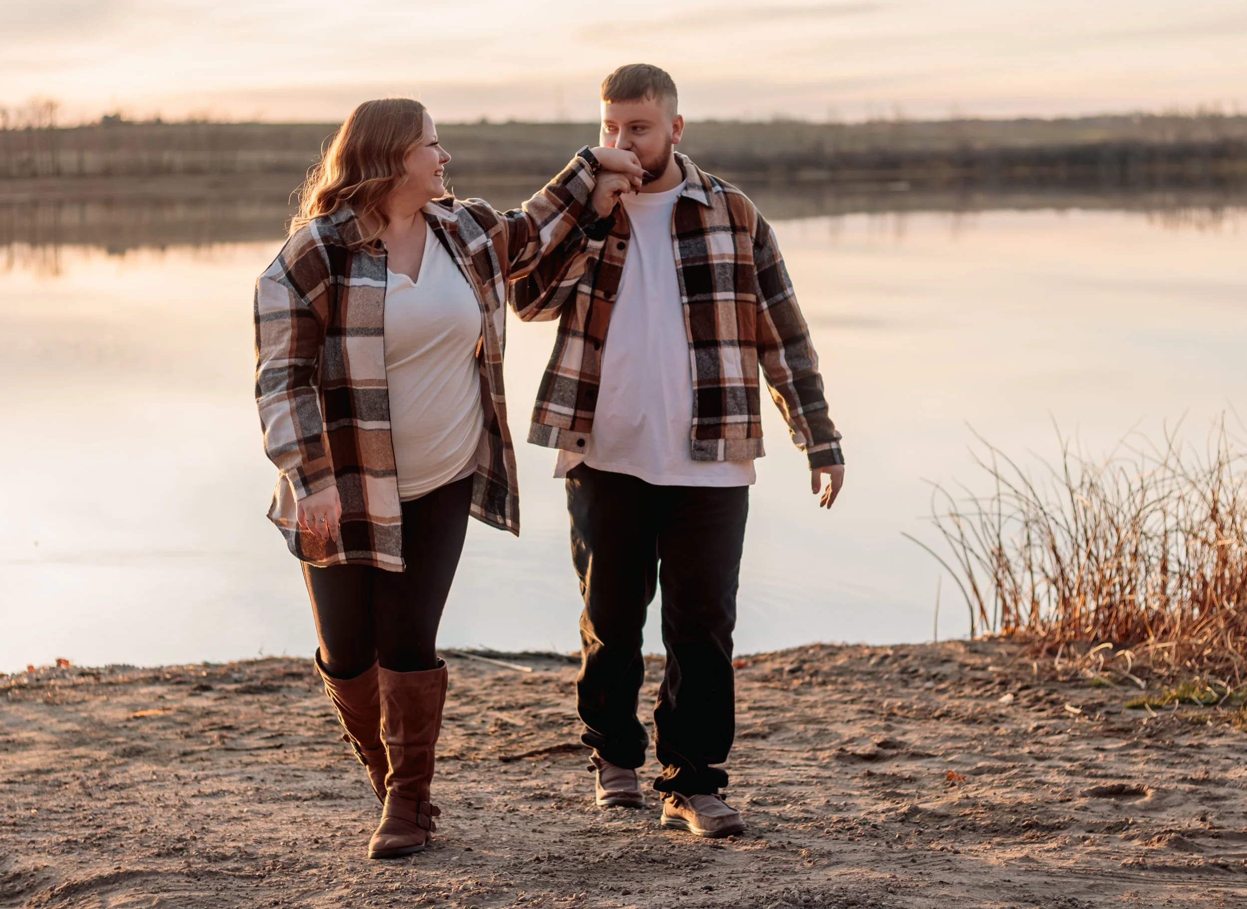 Couple walking beside a calm lake at sunset, wearing plaid jackets and smiling affectionately.
