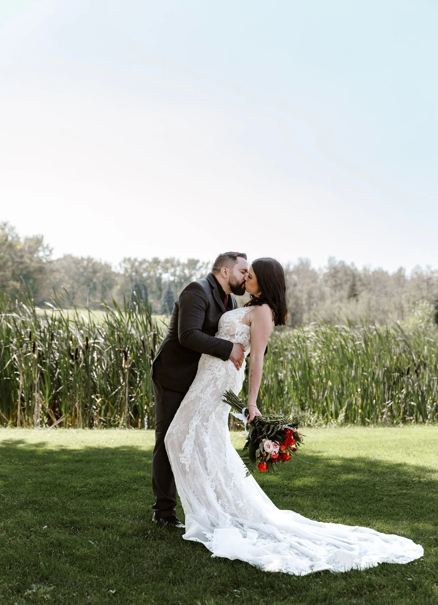 A wedding scene outdoors with a bride and groom, a woman, and a young boy under a large evergreen tree. The bride is holding a bouquet of red flowers, and rose petals are scattered on the grass.