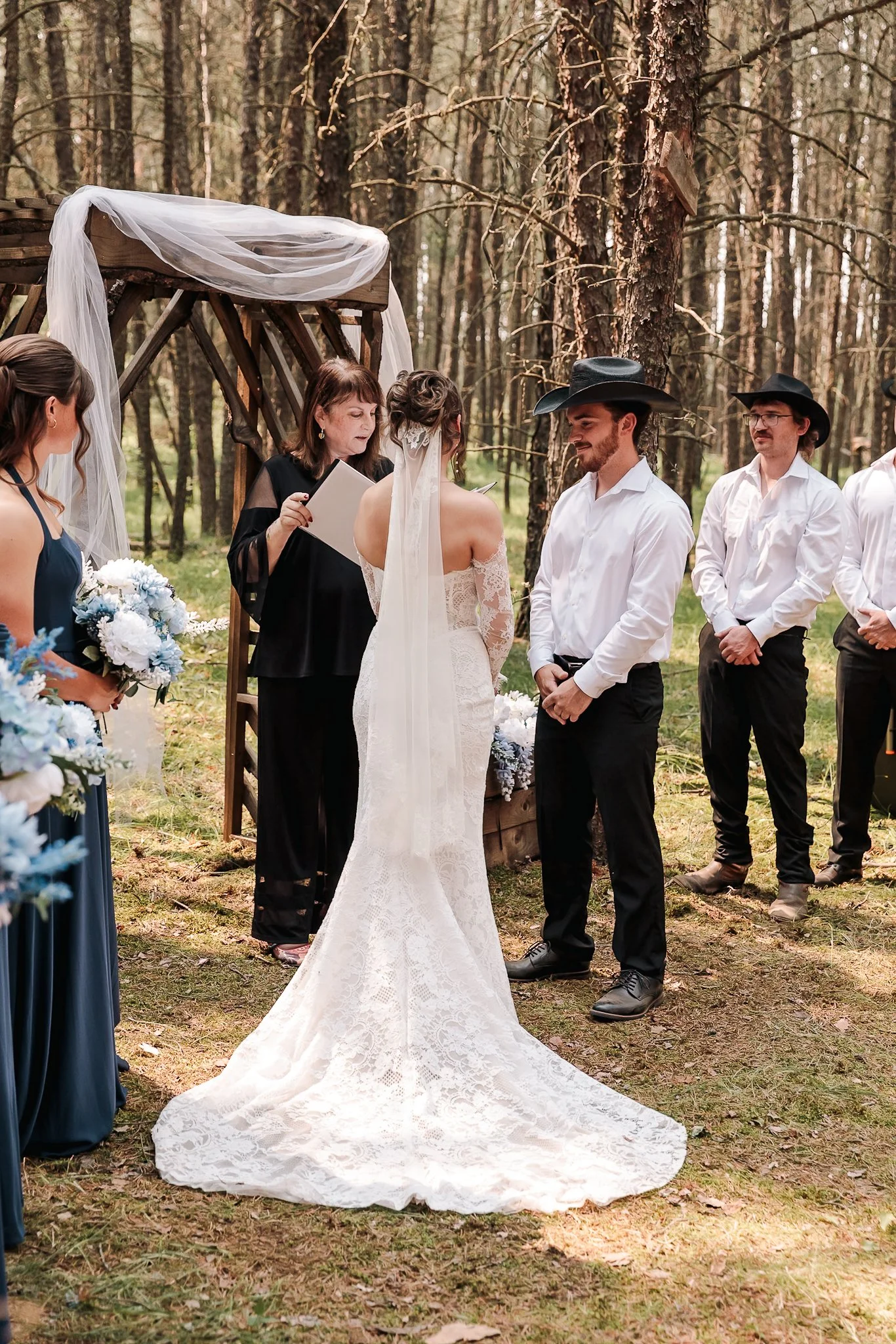 A wedding ceremony taking place outdoors in a forest, featuring a bride in a white lace wedding gown with a long train, a groom in a white shirt and black hat, and an officiant reading from a book. Guests are dressed in formal attire, and there are f