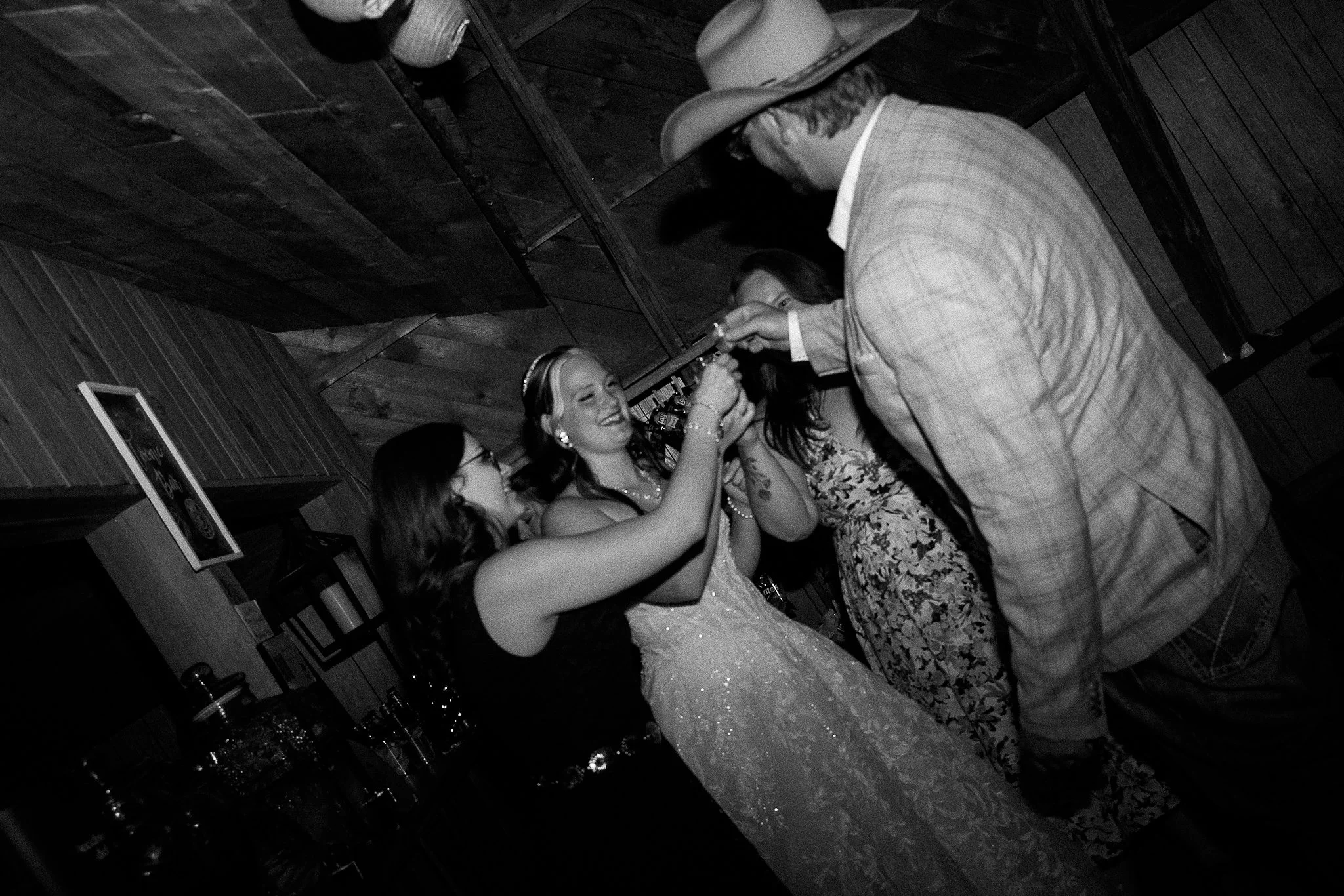 A woman in a wedding dress and a man in a plaid jacket and cowboy hat are dancing together at a party, with other women visible in the background.