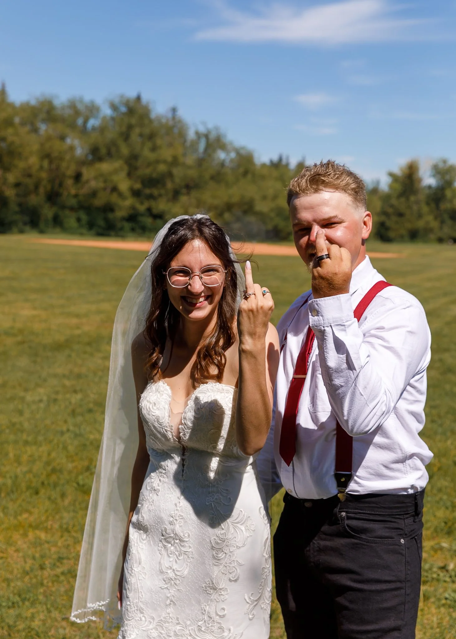 A smiling bride and groom standing outdoors on a sunny day with a grassy field and trees in the background. Both are showing their middle fingers.