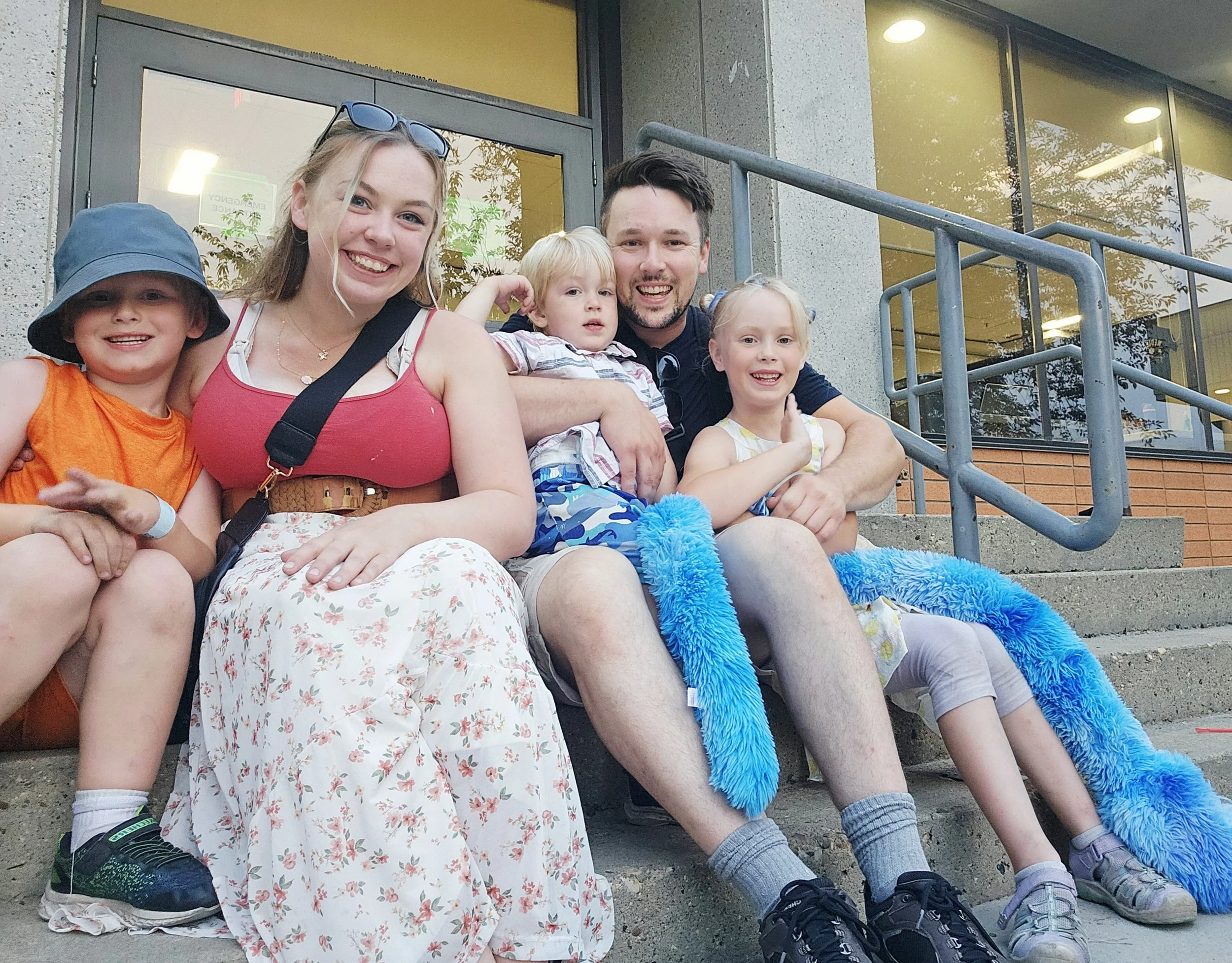 Group of five people, including two children and three adults, sitting on the steps outside a building. They appear happy and relaxed, with some smiling at the camera.