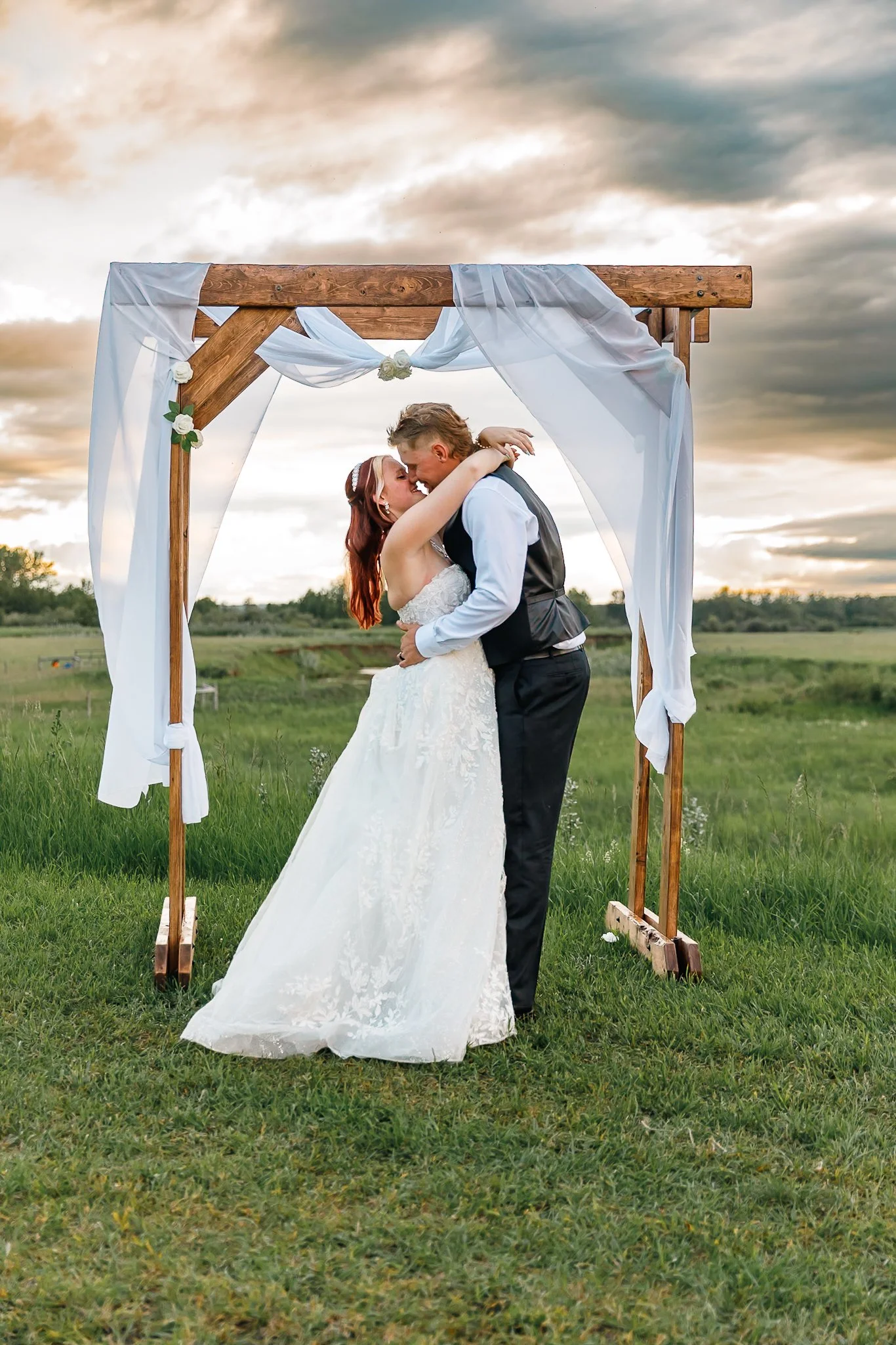 A bride and groom sharing a kiss under a decorated wedding arch outdoors during sunset.