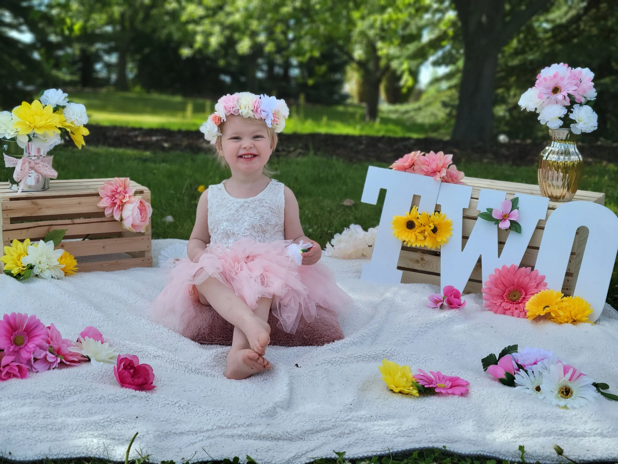 A young girl with blonde hair, smiling, sitting on a white fluffy rug outdoors. She is wearing a white lace top, a pink tulle skirt, and a headband made of colorful artificial flowers. Around her are more artificial flowers, some in wooden crates and vases, with a large white decorative sign that spells "LOVE" in pink and yellow flowers. The background shows green trees and grass, indicating a park or garden setting.