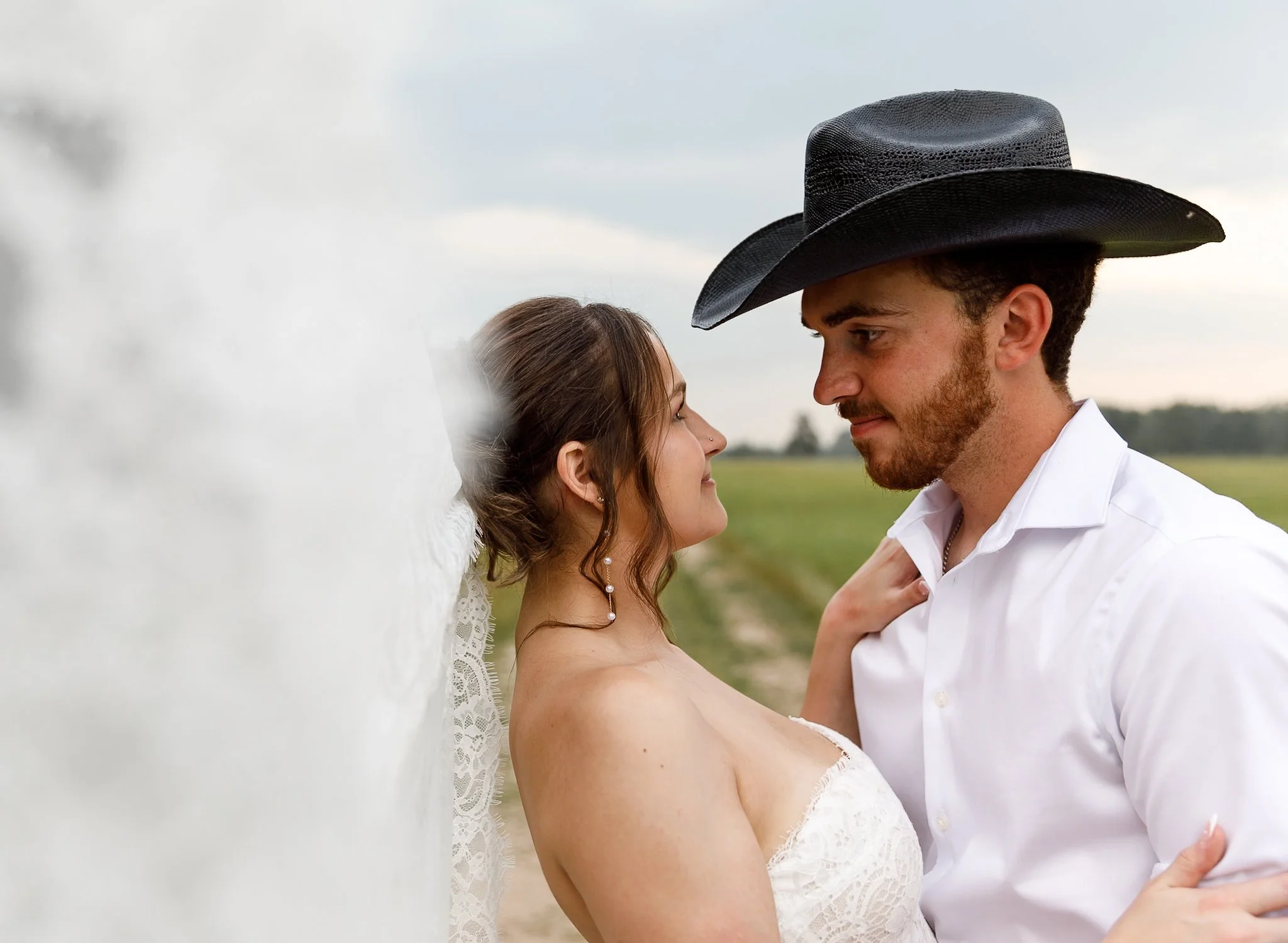 A couple is standing outdoors, facing each other close with their foreheads touching, with a woman in a white lace dress and a man in a white shirt and a black cowboy hat. They are smiling softly at each other, with a green field and cloudy sky in th