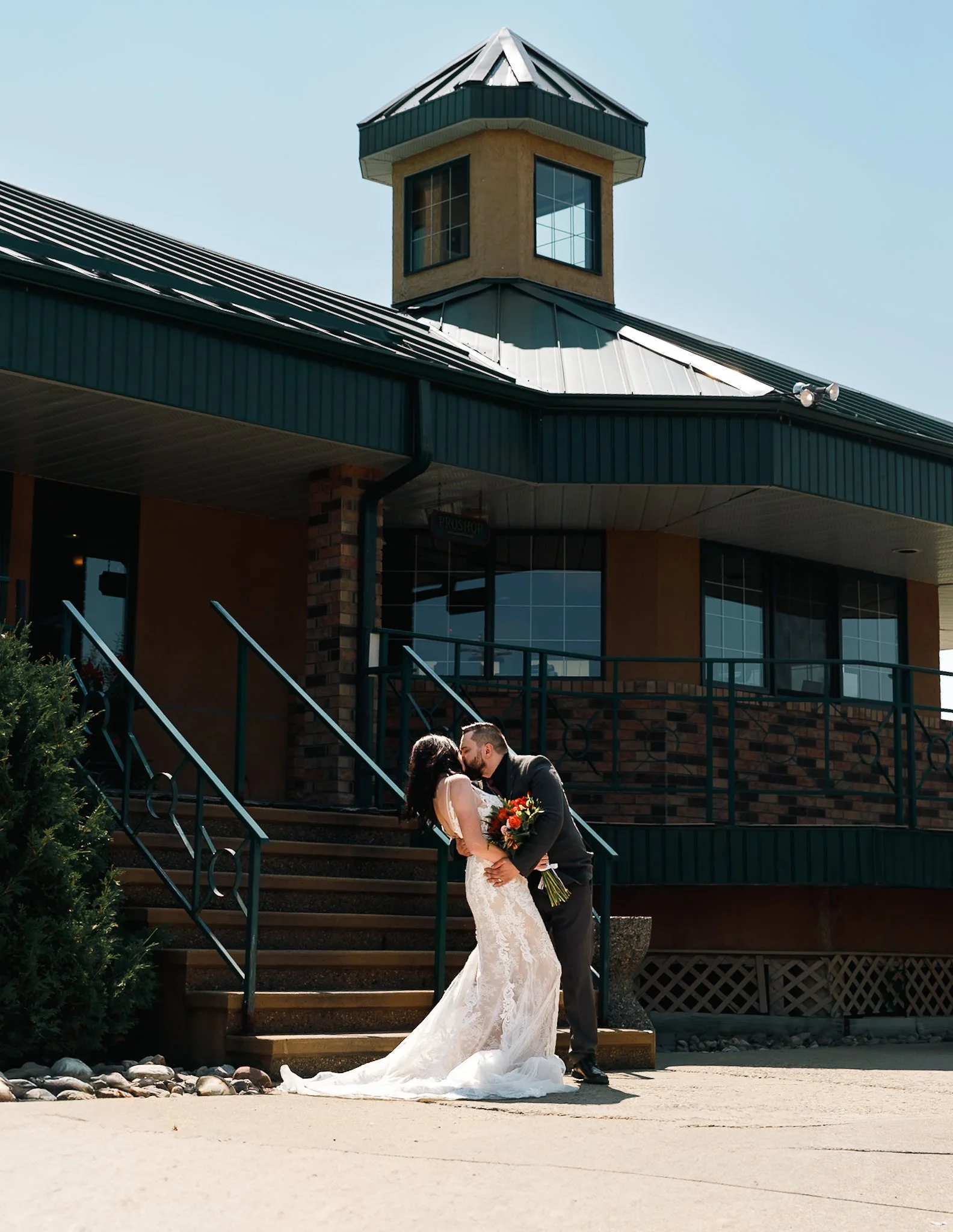 A bride and groom share a kiss outside a building with a turret. The bride is wearing a white lace wedding gown and holding a bouquet. The groom is in a suit. They are standing on stairs with rocks in the foreground.