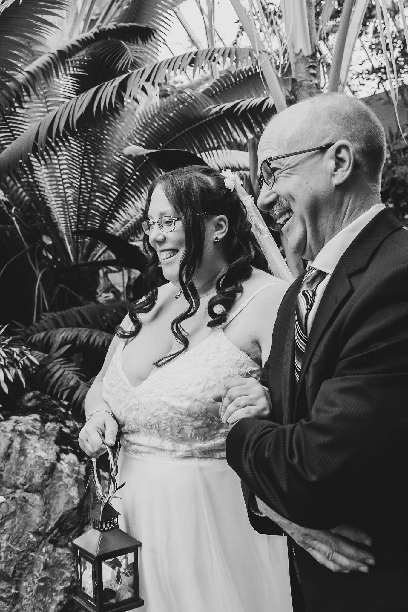 A black and white photo of a bride and groom holding hands, smiling, surrounded by tropical plants and lanterns.