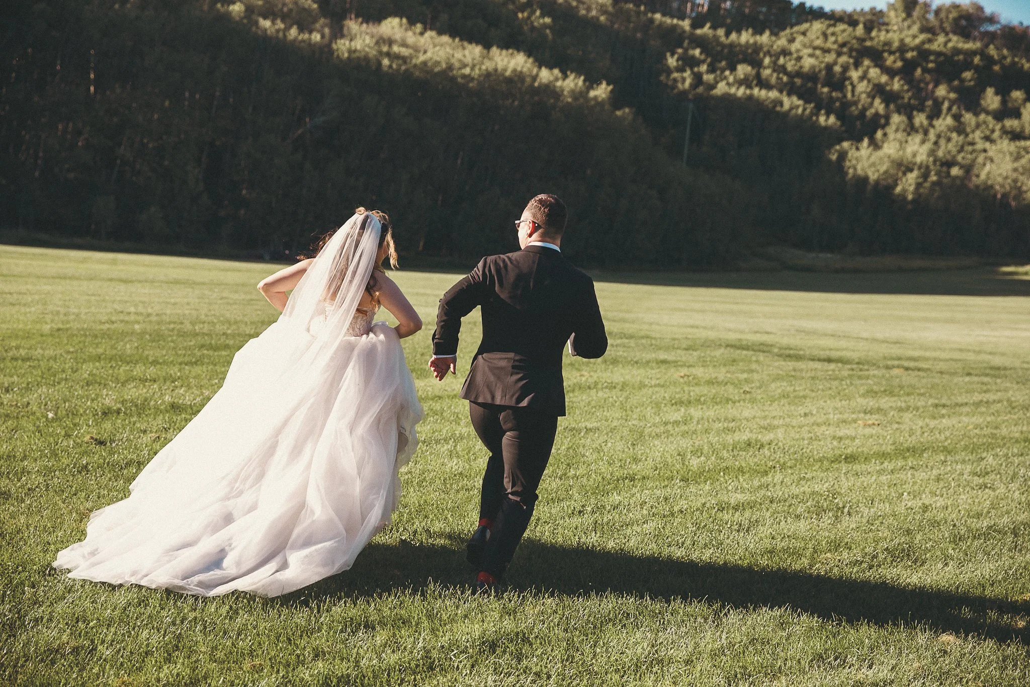 A bride in a white wedding gown and veil and a groom in a black suit running across a green field with trees in the background.