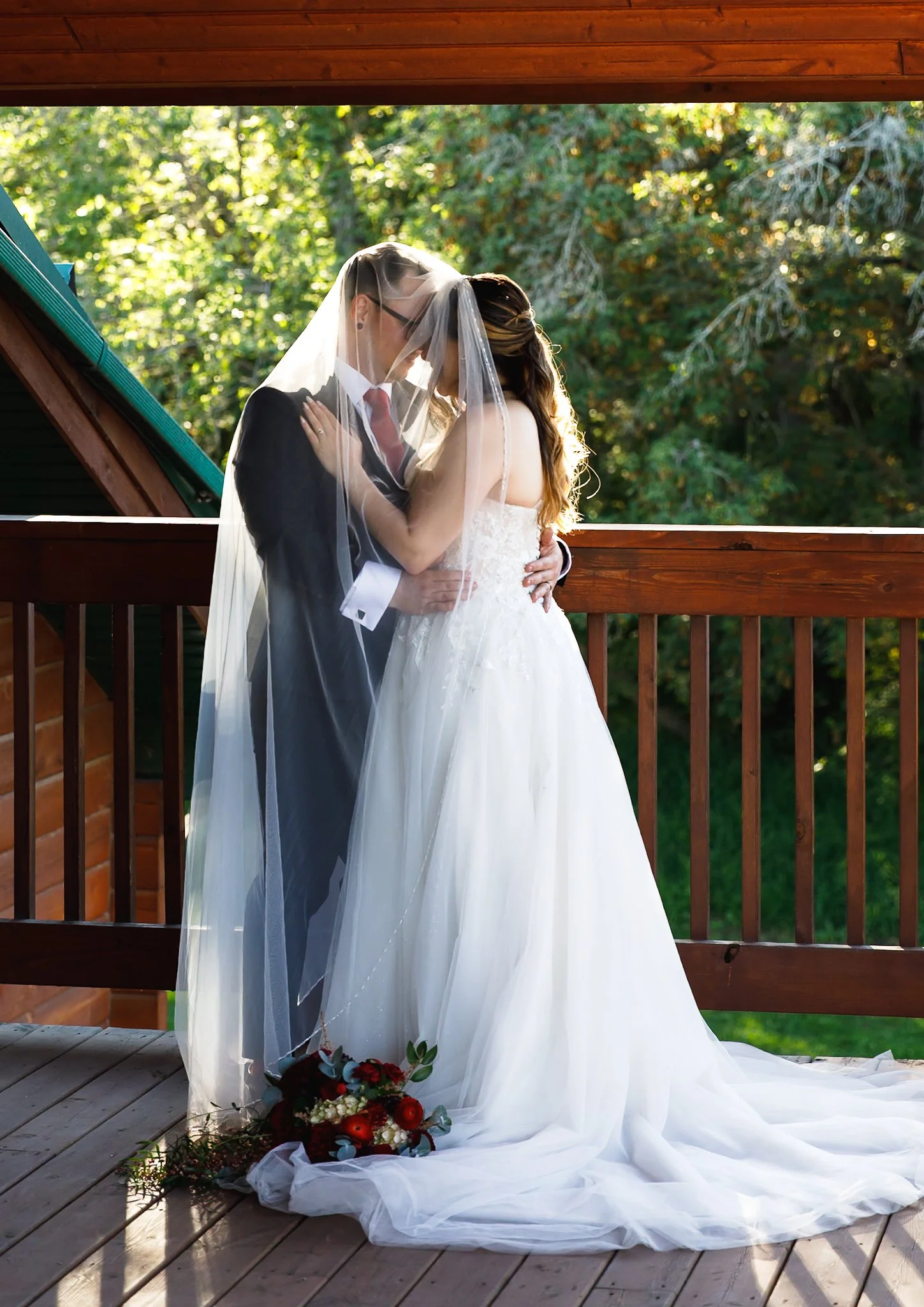 A bride and groom sharing a kiss under a veil on a wooden deck, with a bouquet of red and white flowers on the ground and lush green trees in the background.