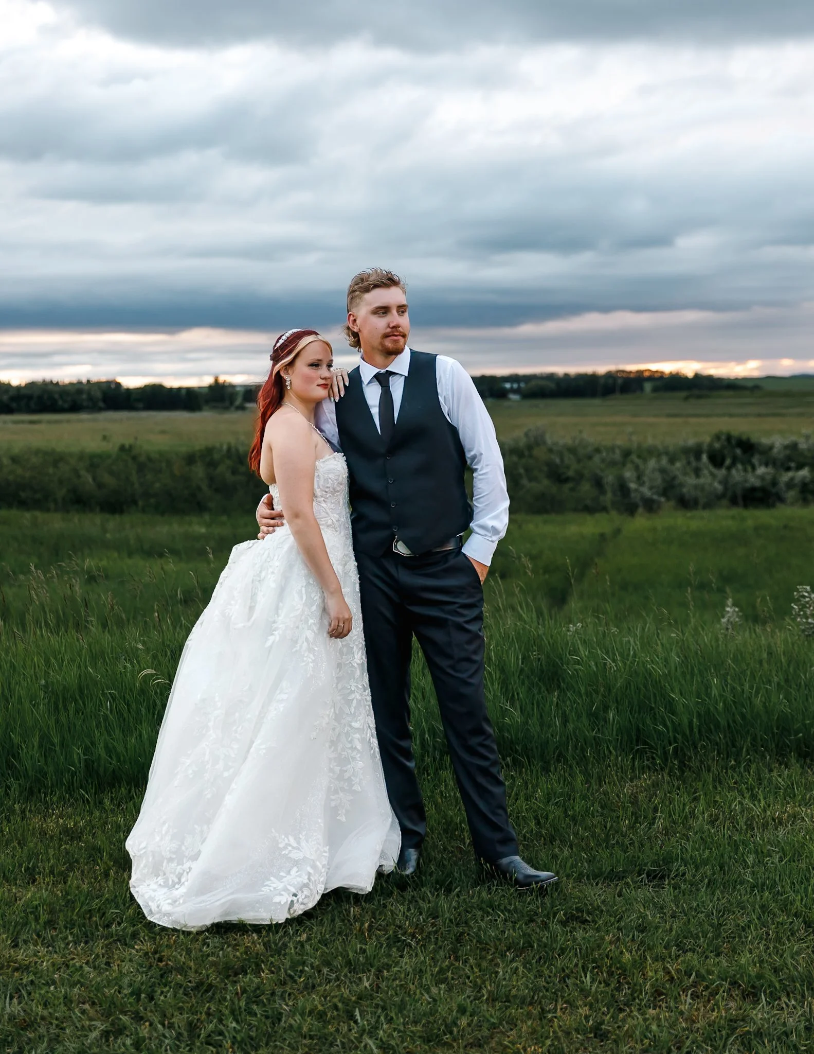 A couple dressed in wedding attire standing in a green field under a cloudy sky at sunset.