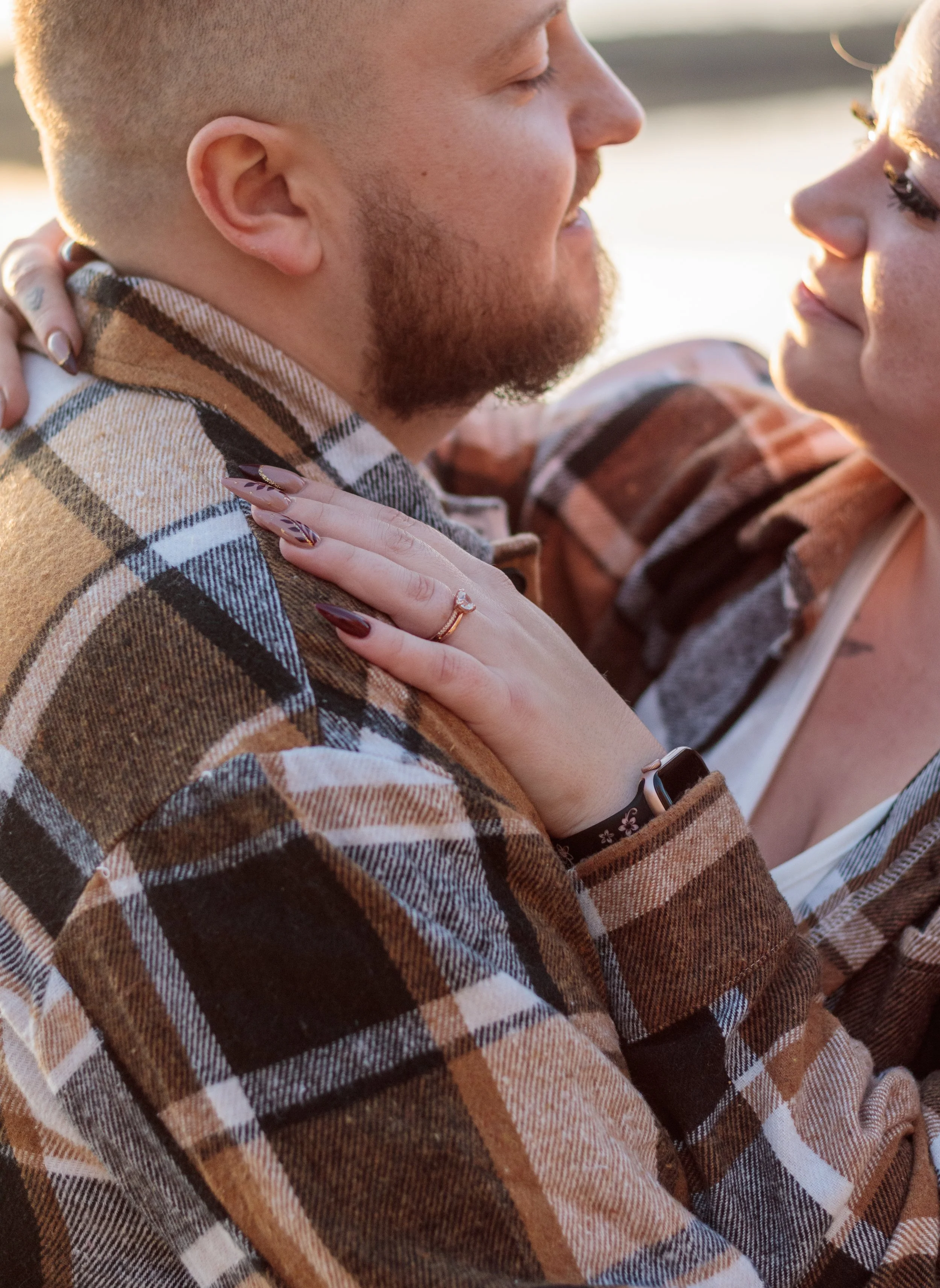 Close-up of a smiling couple embracing outdoors in warm sunlight, with the woman’s hands resting on the bearded man’s plaid jacket.
