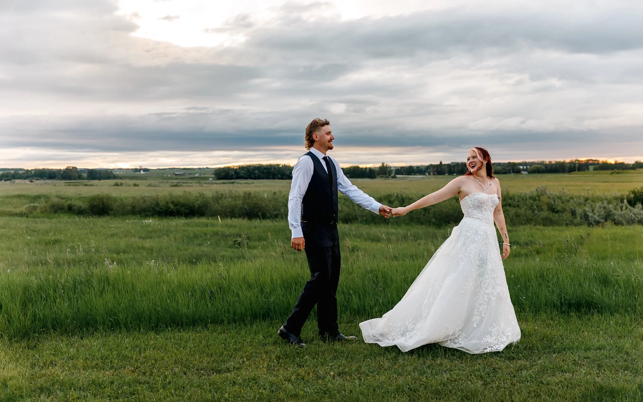 A newlywed couple holding hands and smiling in a grassy field with a cloudy sky and sunset in the background.
